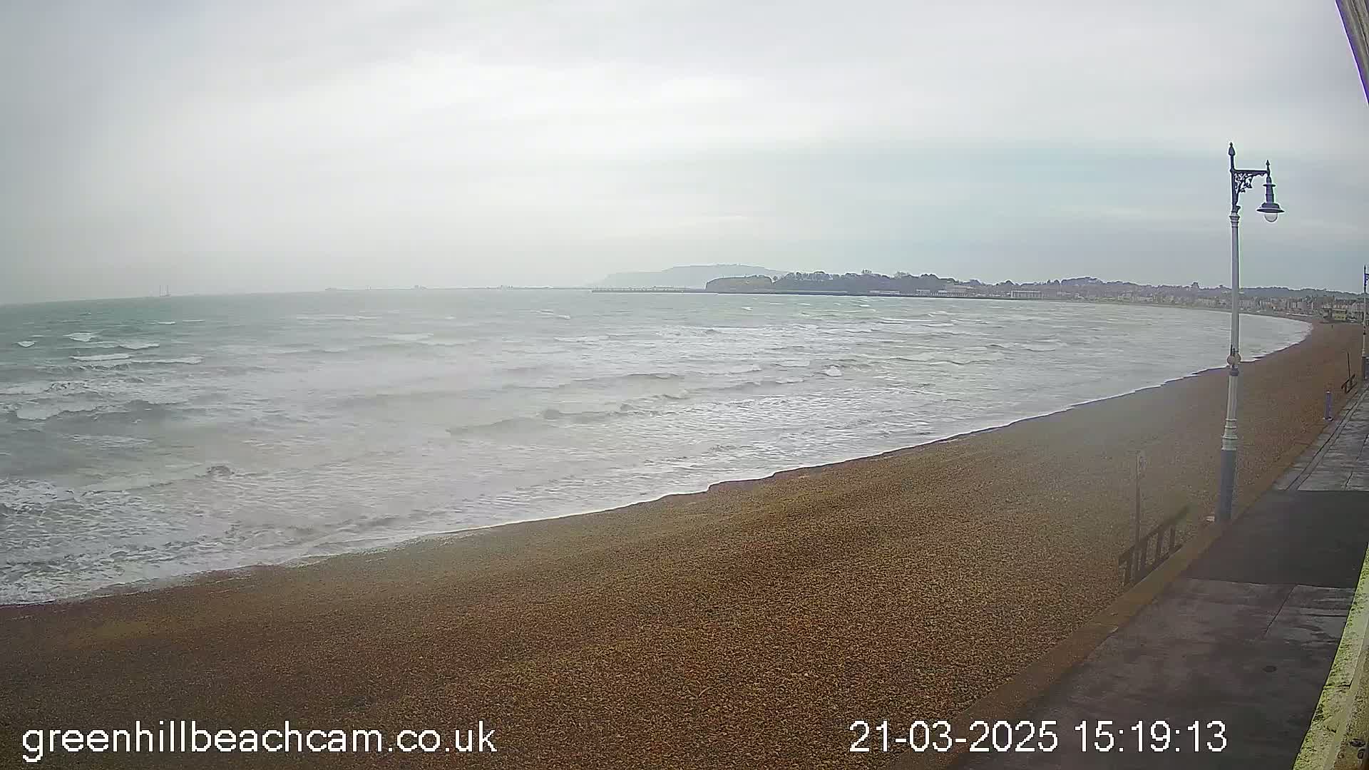 A pebble beach meets a choppy sea under an overcast sky, with a distant shoreline visible.