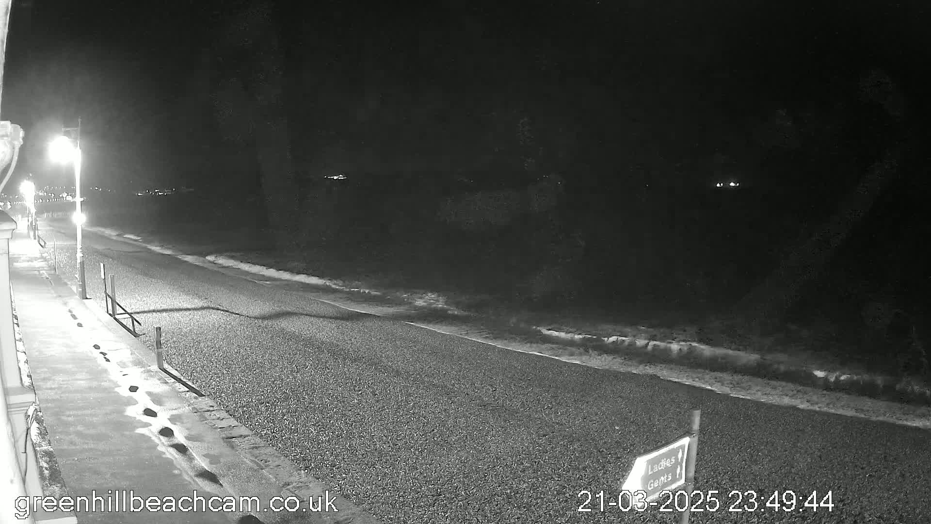 A nighttime, black-and-white view of a pebble beach with gentle waves lapping the shore, illuminated by a single streetlight.