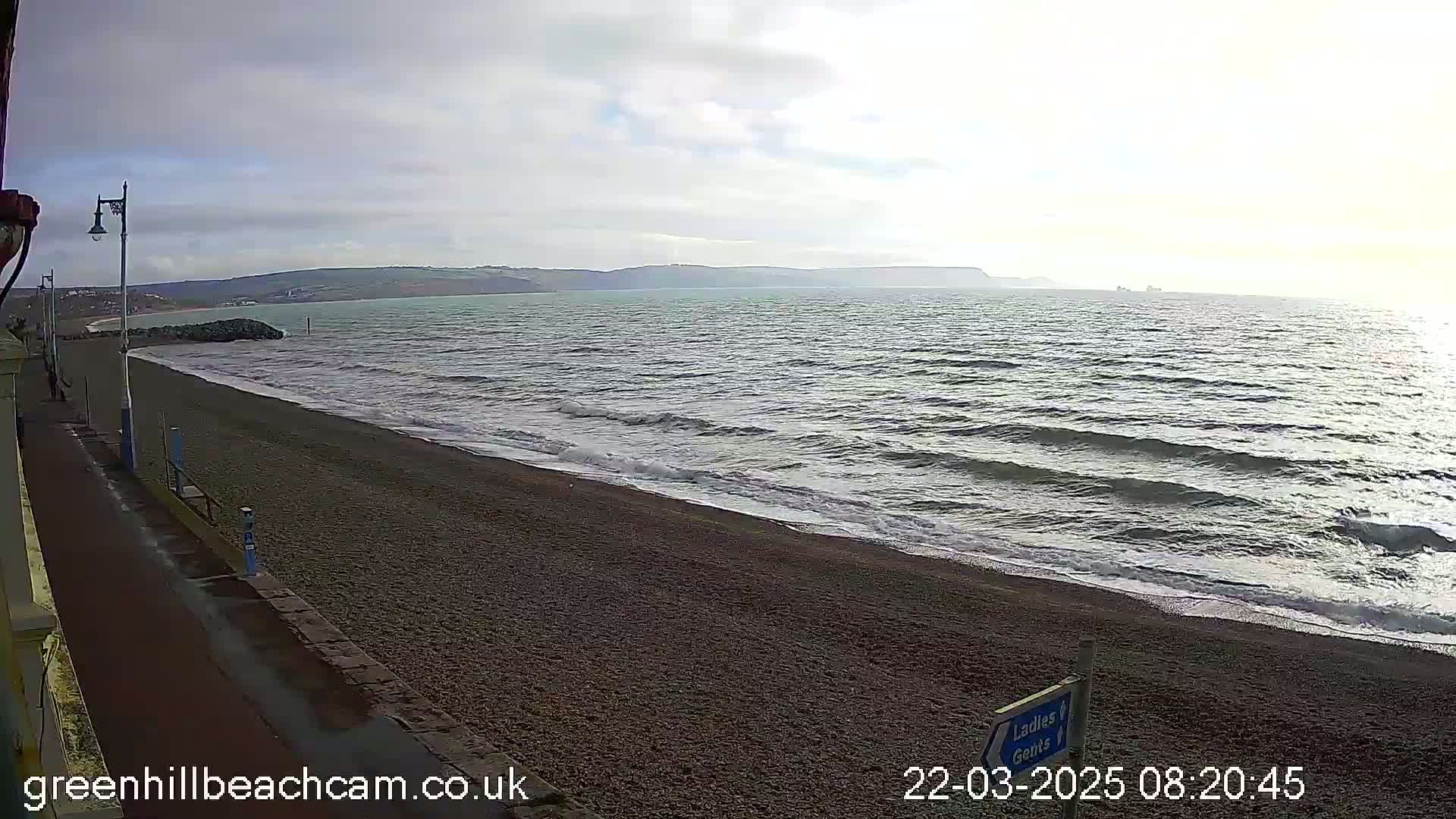 A pebble beach with gentle waves rolls onto the shore under a mostly cloudy sky, with hills visible in the distance.