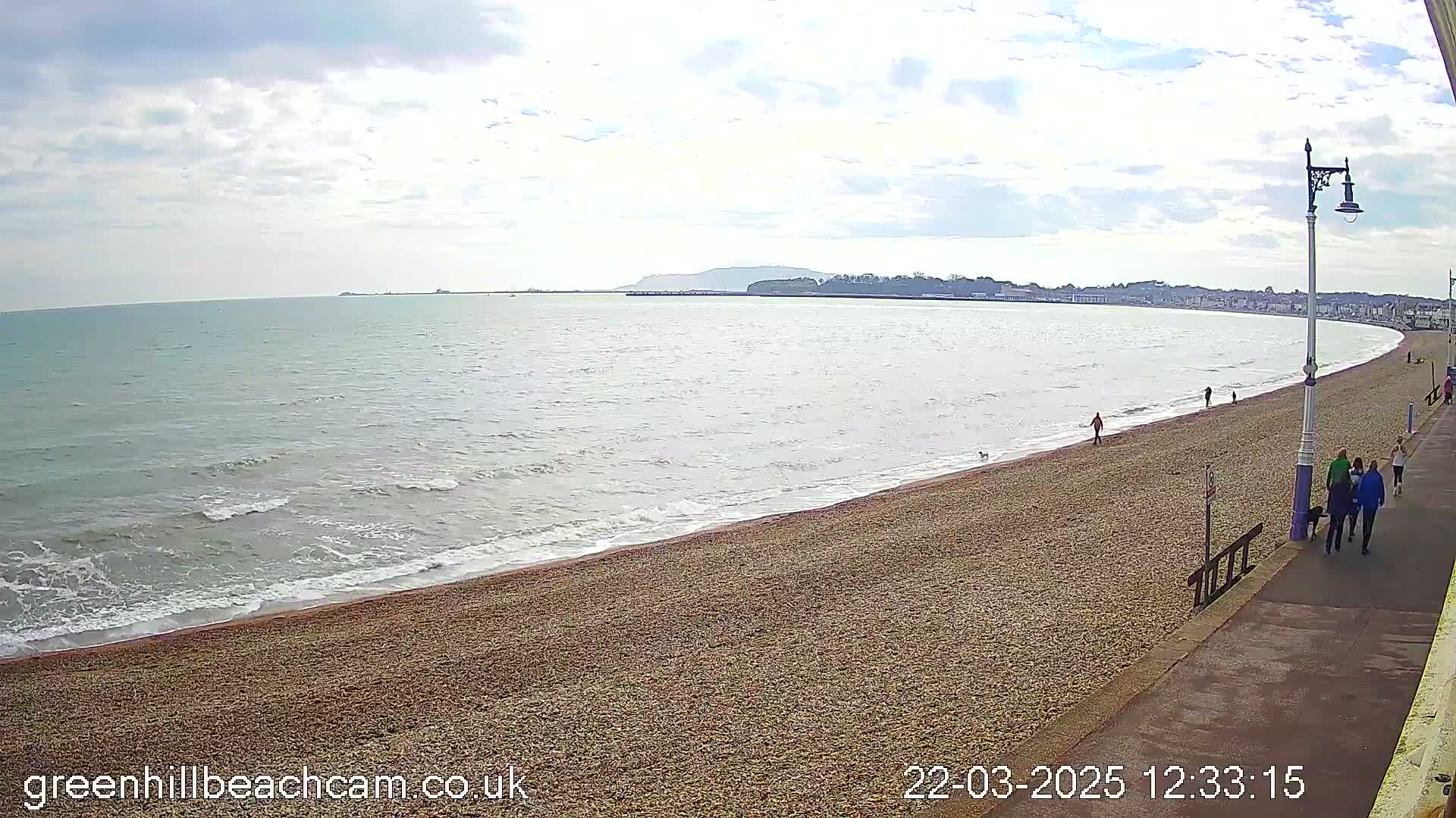 A wide, sandy beach curves gently along a calm sea under a mostly cloudy sky, with a few people walking along the shore and promenade.