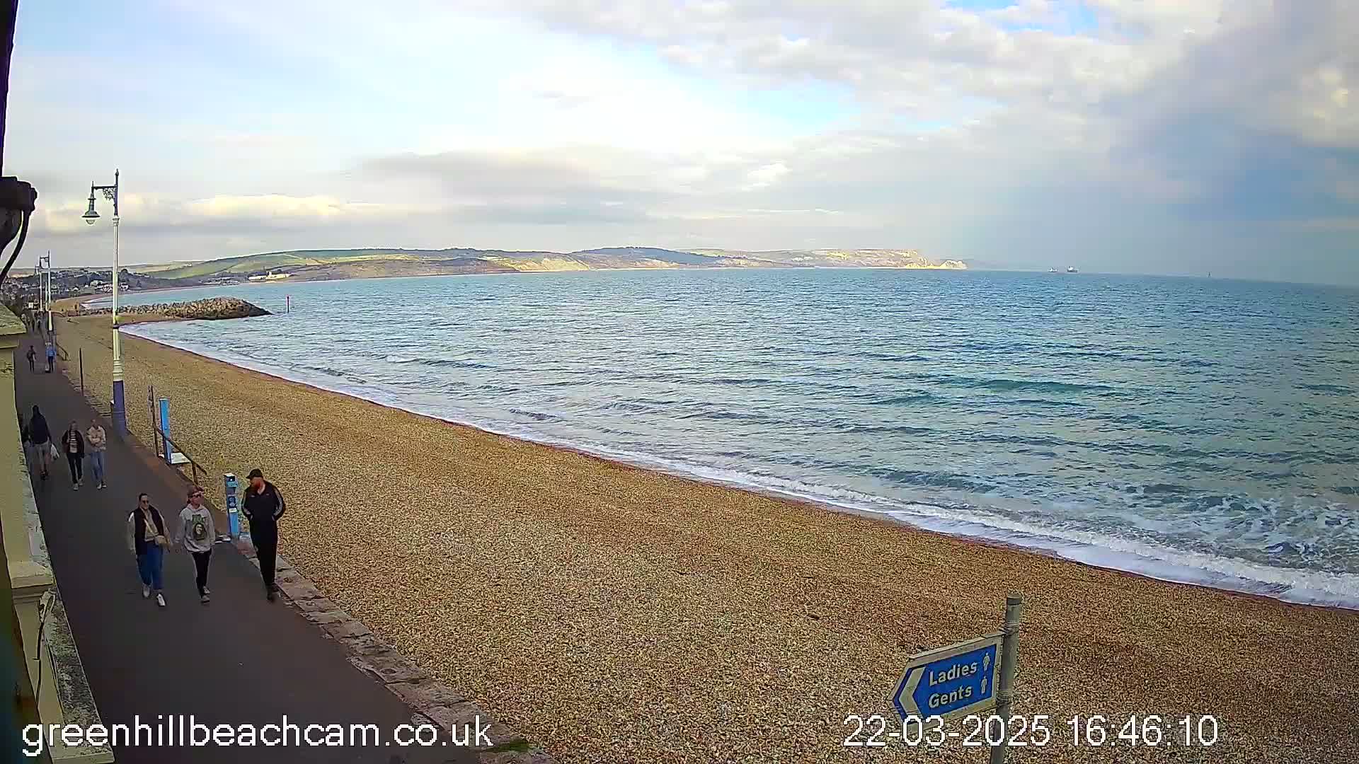 A pebble beach with several people walking along a paved path, overlooking a calm sea under a partly cloudy sky.