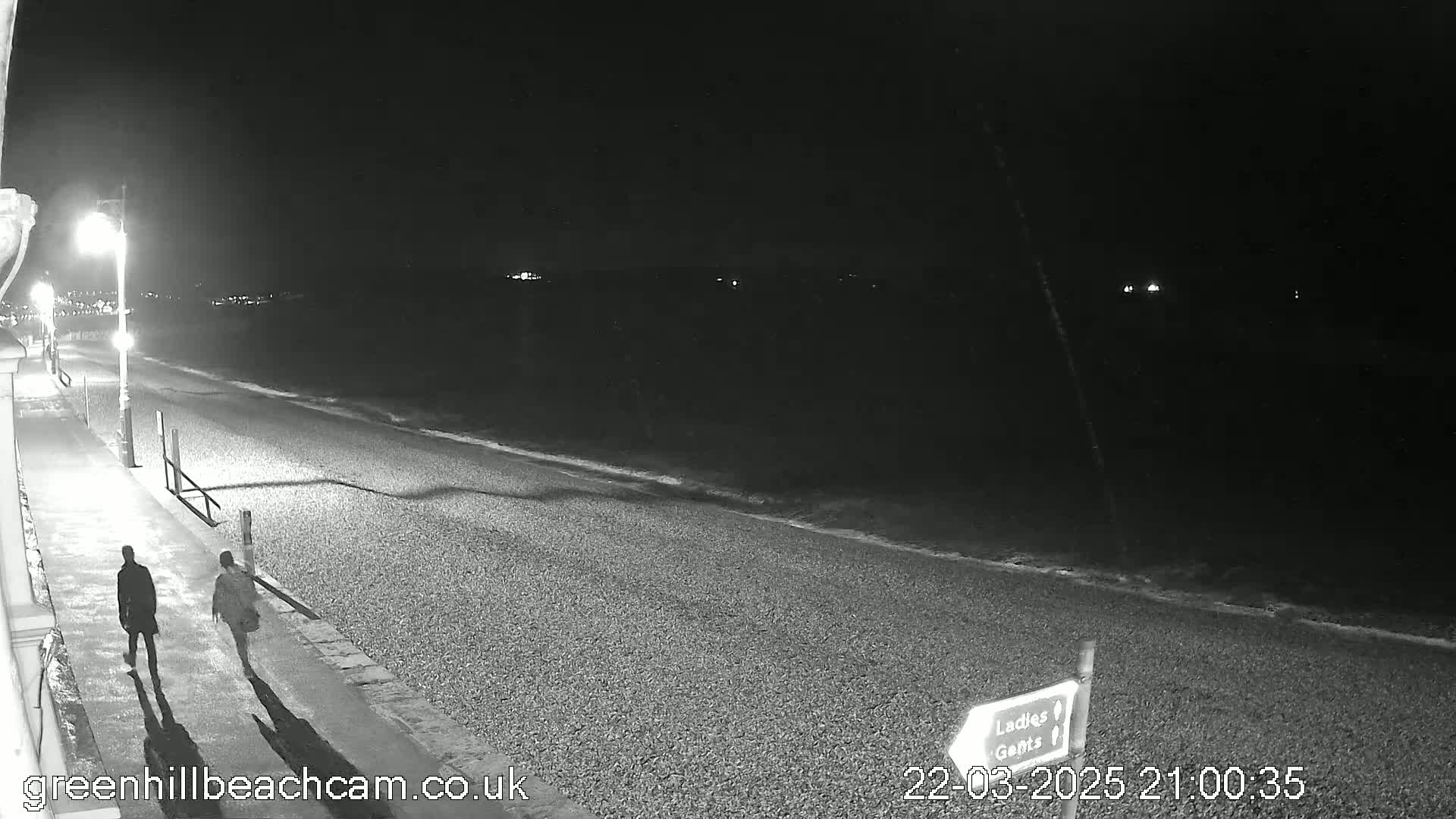 A nighttime black and white view shows two people walking along a pebbled beach path next to a dark ocean under a clear sky.