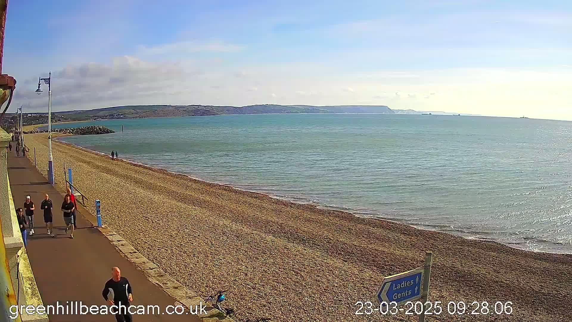 A pebbled beach with several people walking along a paved path beside it, overlooking calm blue water and distant hills under a mostly sunny sky.