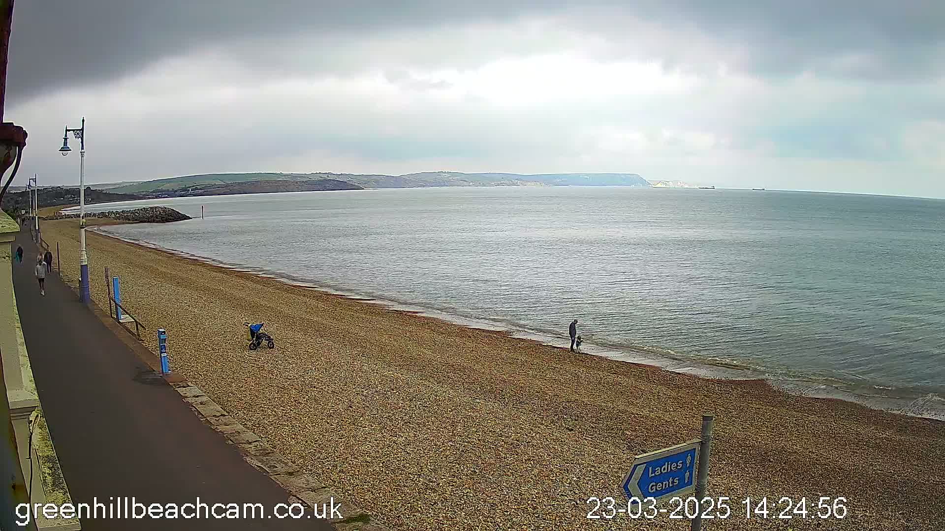 A pebble beach with a few people walking along the shore and a paved walkway next to it, under an overcast sky, overlooks a calm sea with distant hills.