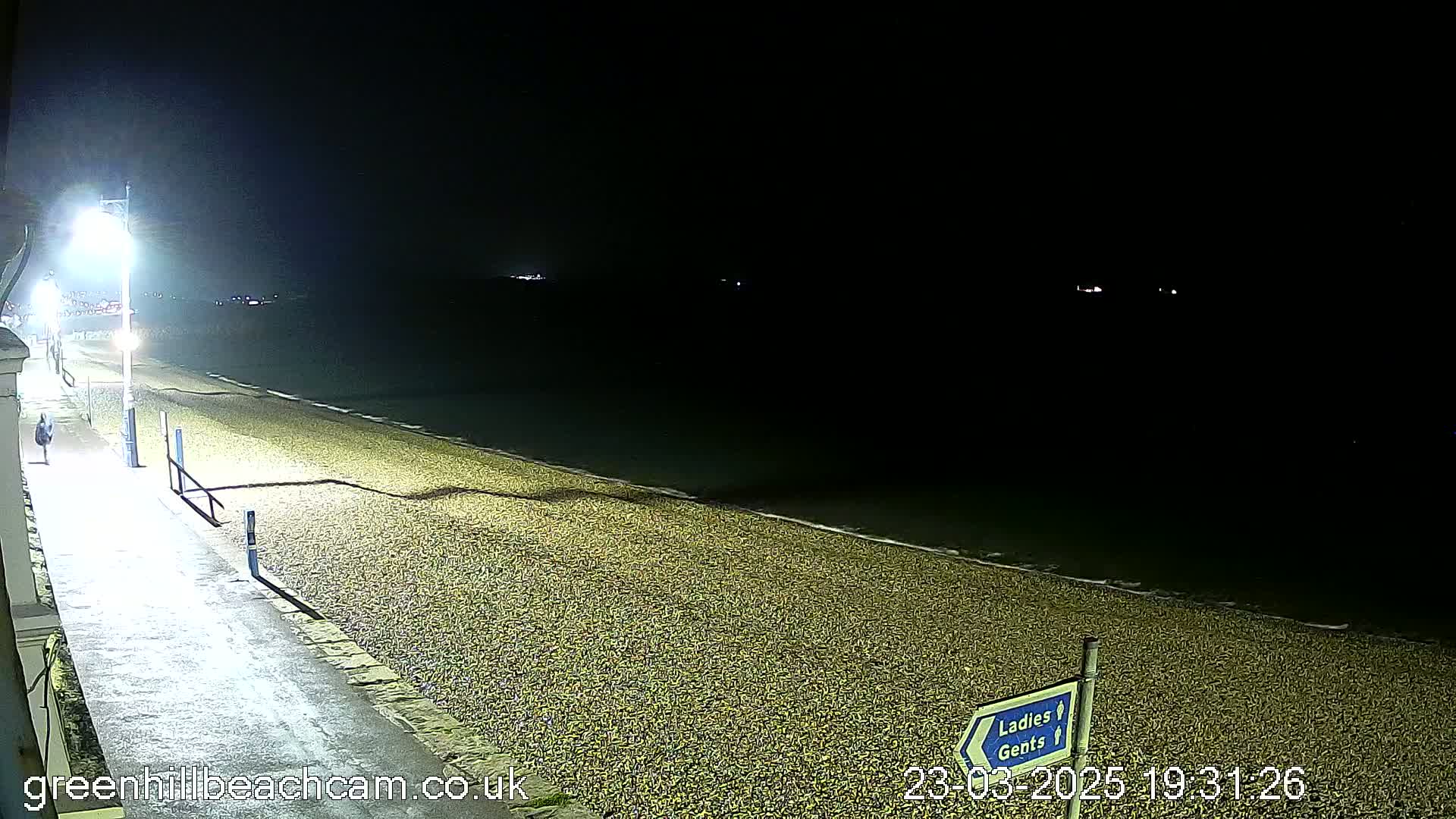 A nighttime view of a pebble beach with a paved walkway and a single person walking along it, under partly cloudy conditions.
