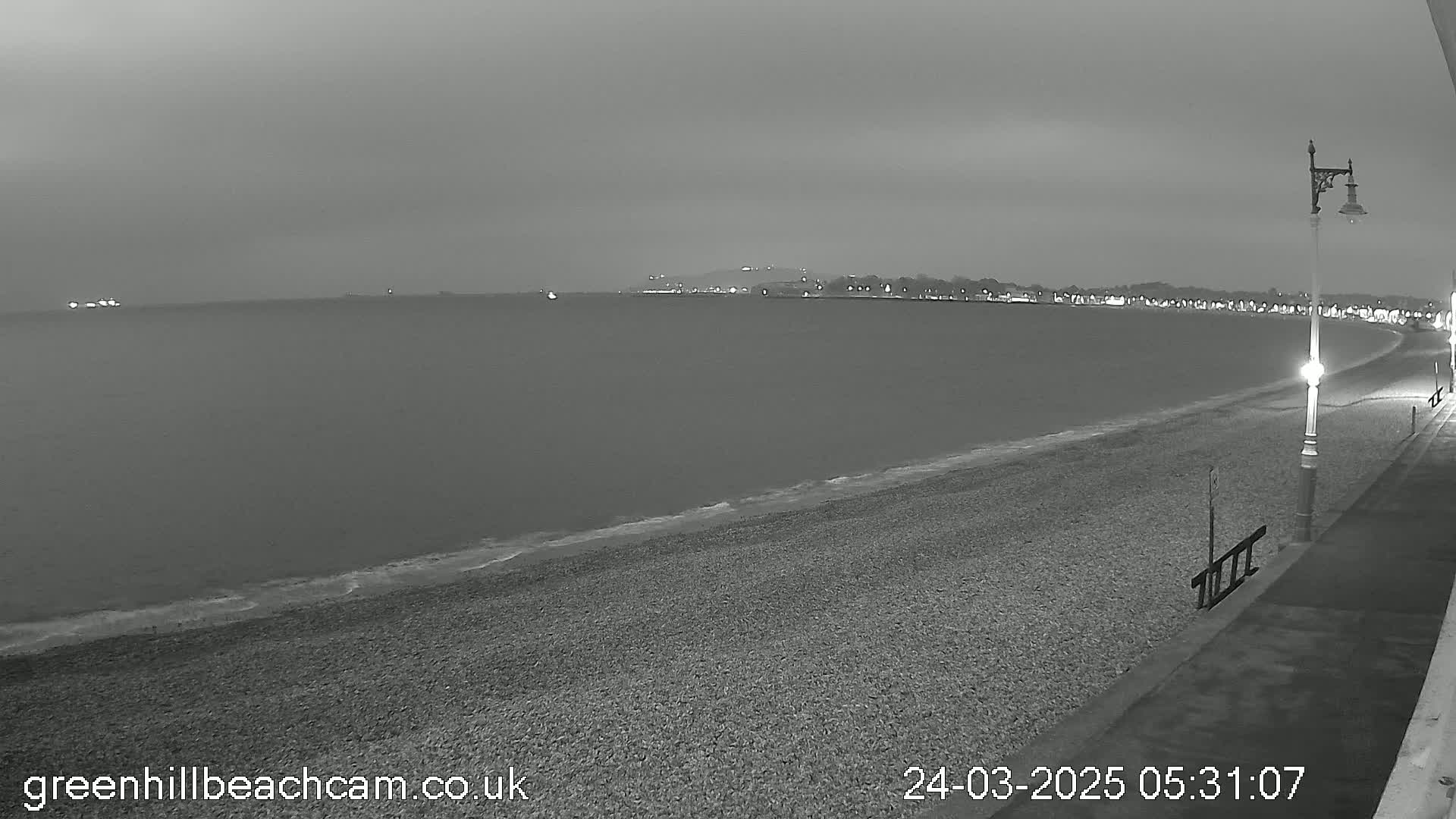 A dark, overcast night scene shows a pebble beach curving gently toward a calm sea under a gray sky, with lights visible in the distance along a shoreline.
