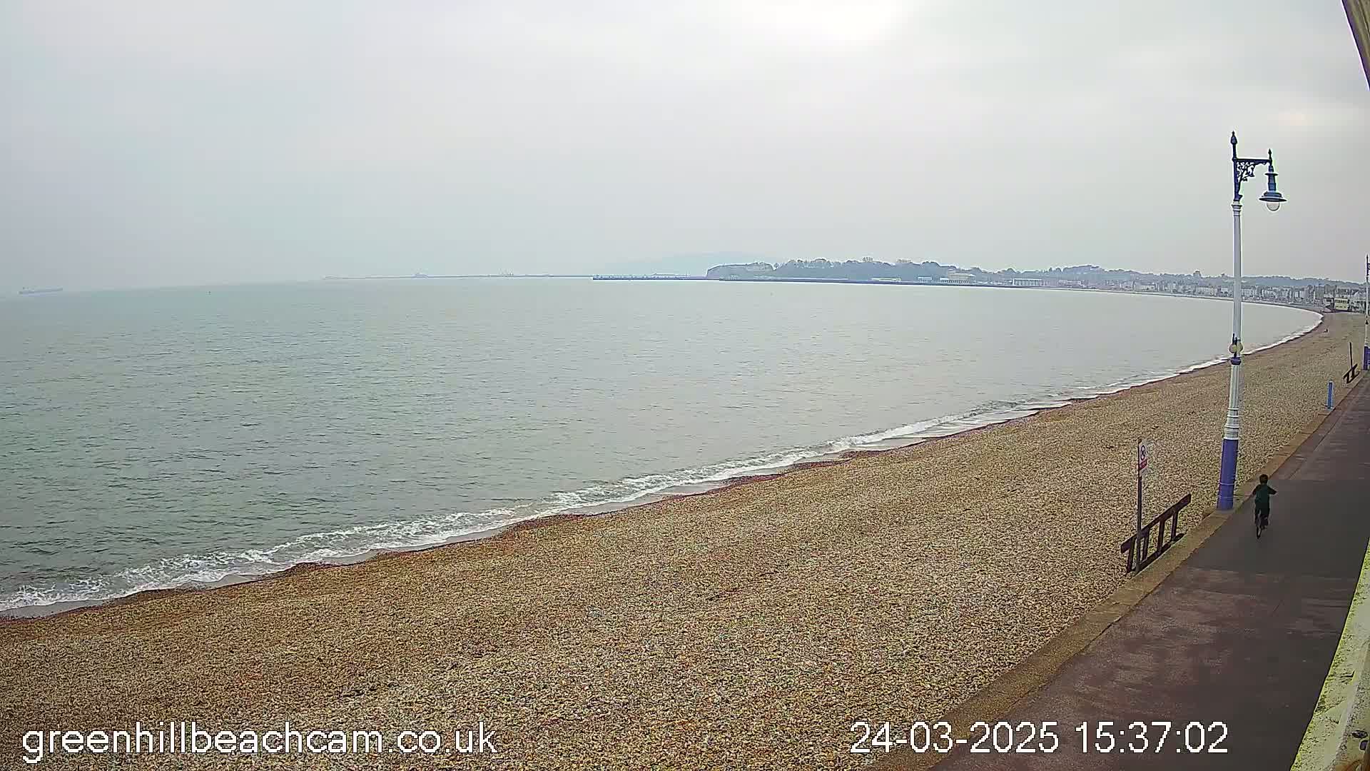 A person runs along a paved walkway next to a pebble beach and calm, gray sea under an overcast sky.
