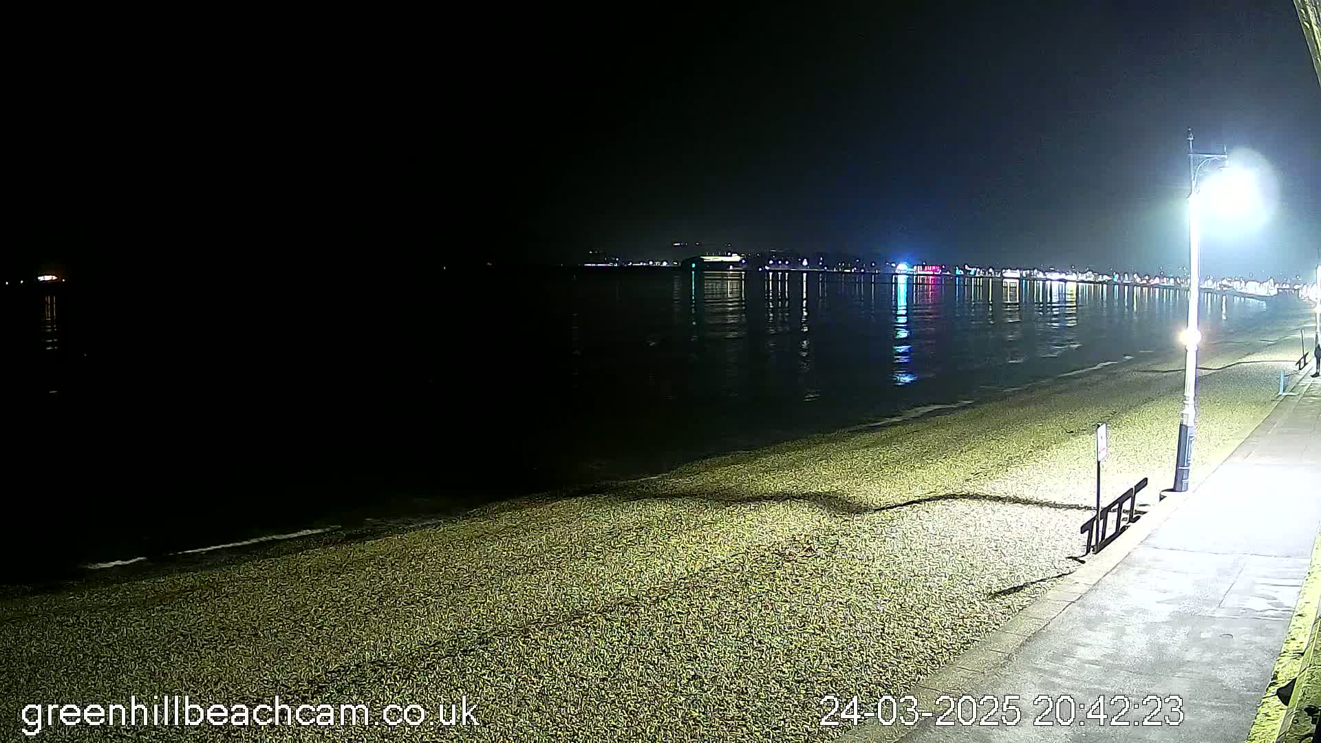 A dark, nighttime beach scene shows a pebble shoreline, calm water reflecting distant lights, and a paved walkway with streetlights.