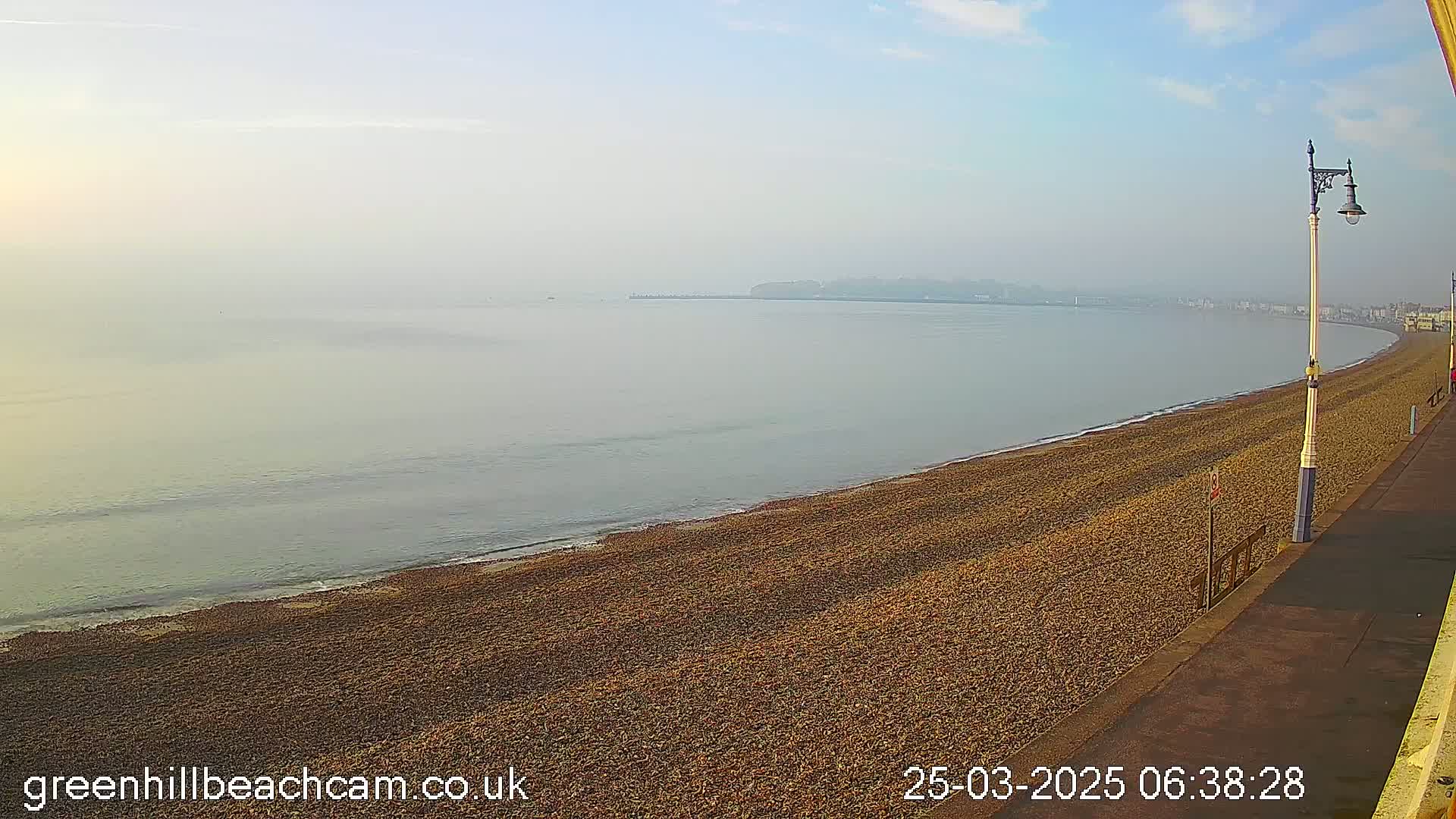 A hazy, calm morning scene shows a long, gently curving pebble beach meeting a calm sea under a clear, light blue sky.