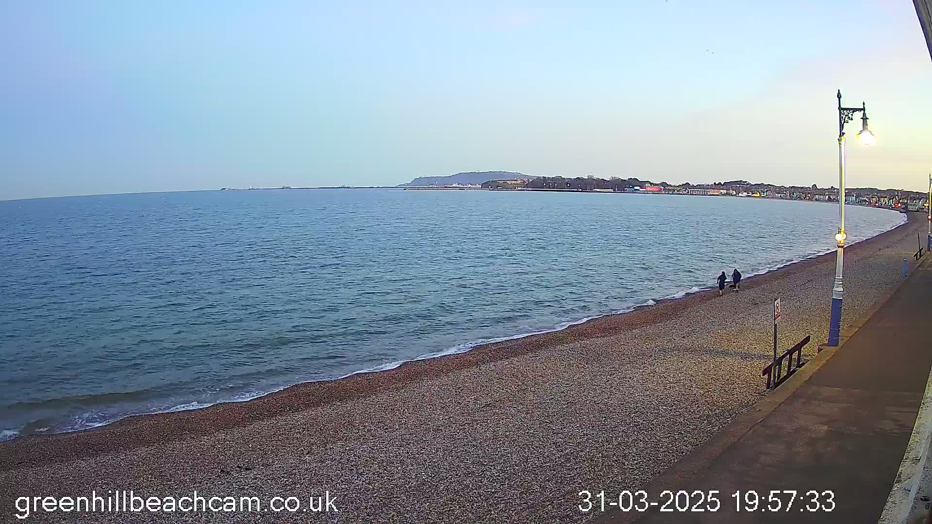 A calm sea laps a pebble beach under a clear, light-blue sky, with two people and a dog walking along the shore near a promenade.