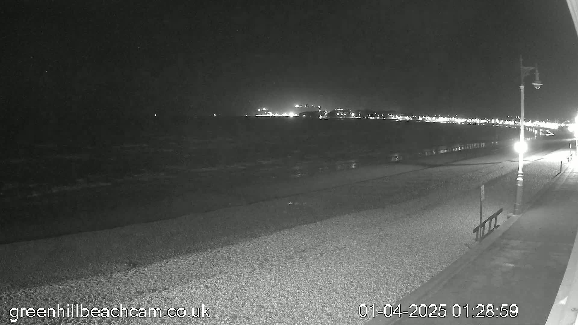 A nighttime view of a pebble beach with a calm sea and a distant town illuminated, under dark skies.