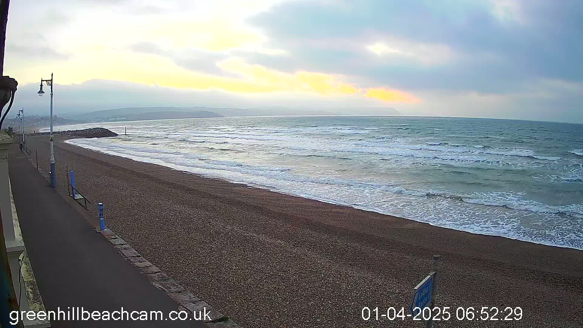 A pebble beach with moderate waves under a partly cloudy sky shows a hint of sunrise.