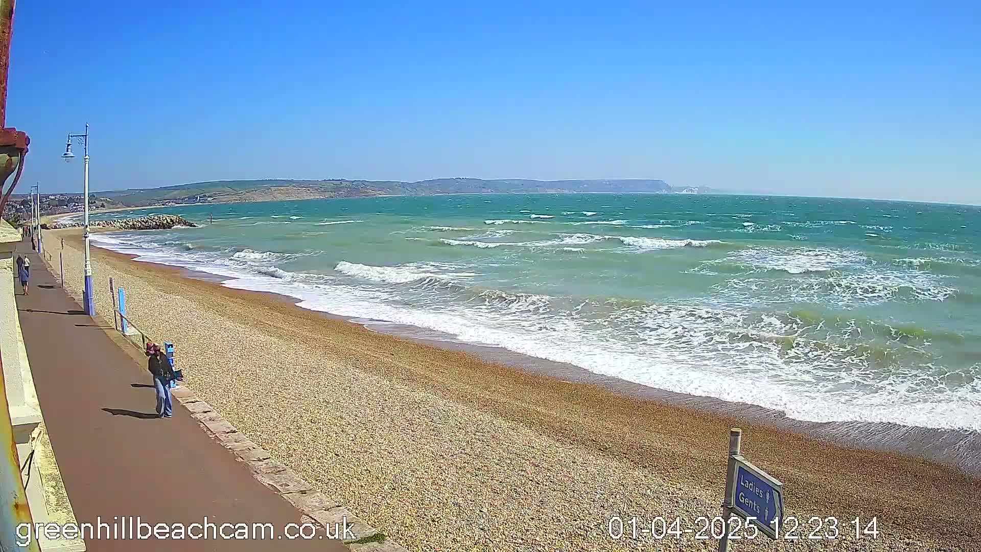 A windy, sunny day at a pebble beach shows moderately sized waves crashing on the shore with a few people walking along a paved path next to the beach.