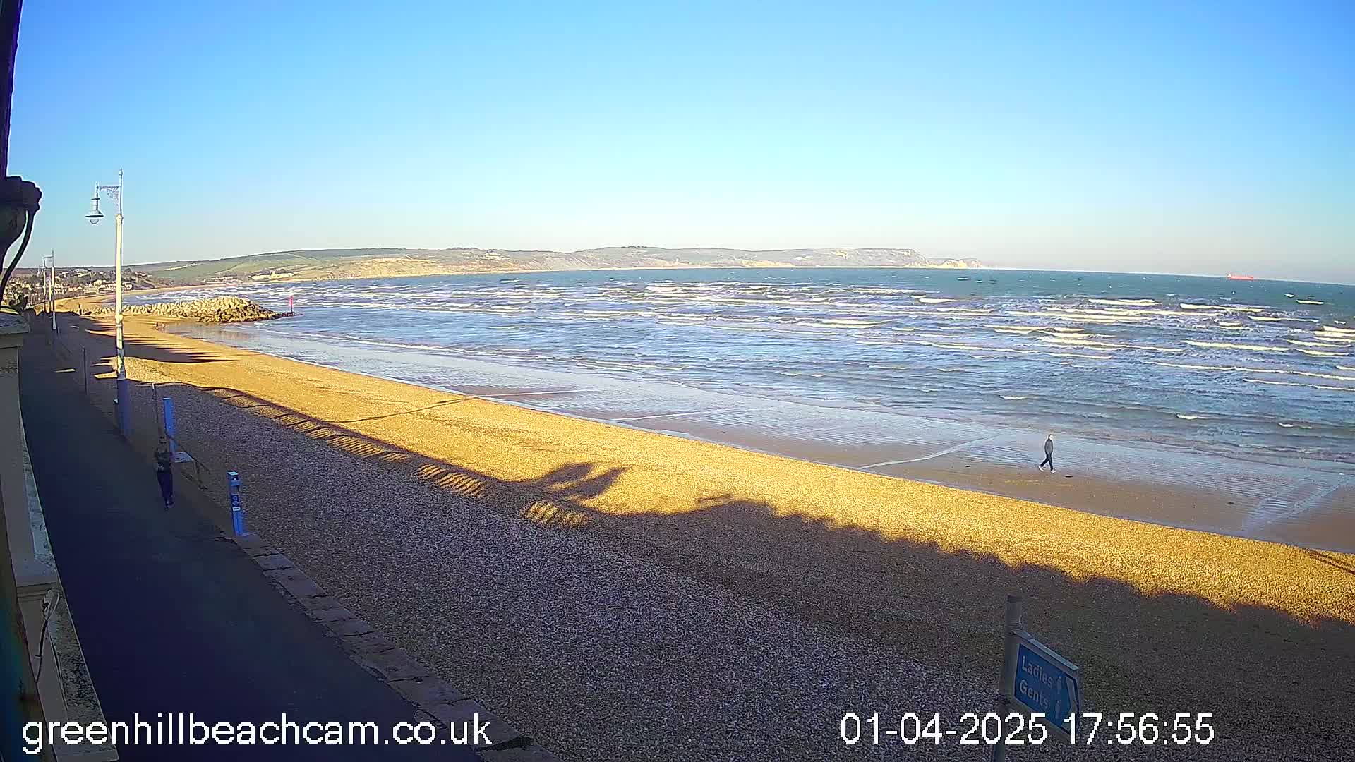 A sandy beach with gentle waves under a clear blue sky, a person walking along the shore, and shadows cast on the sand.