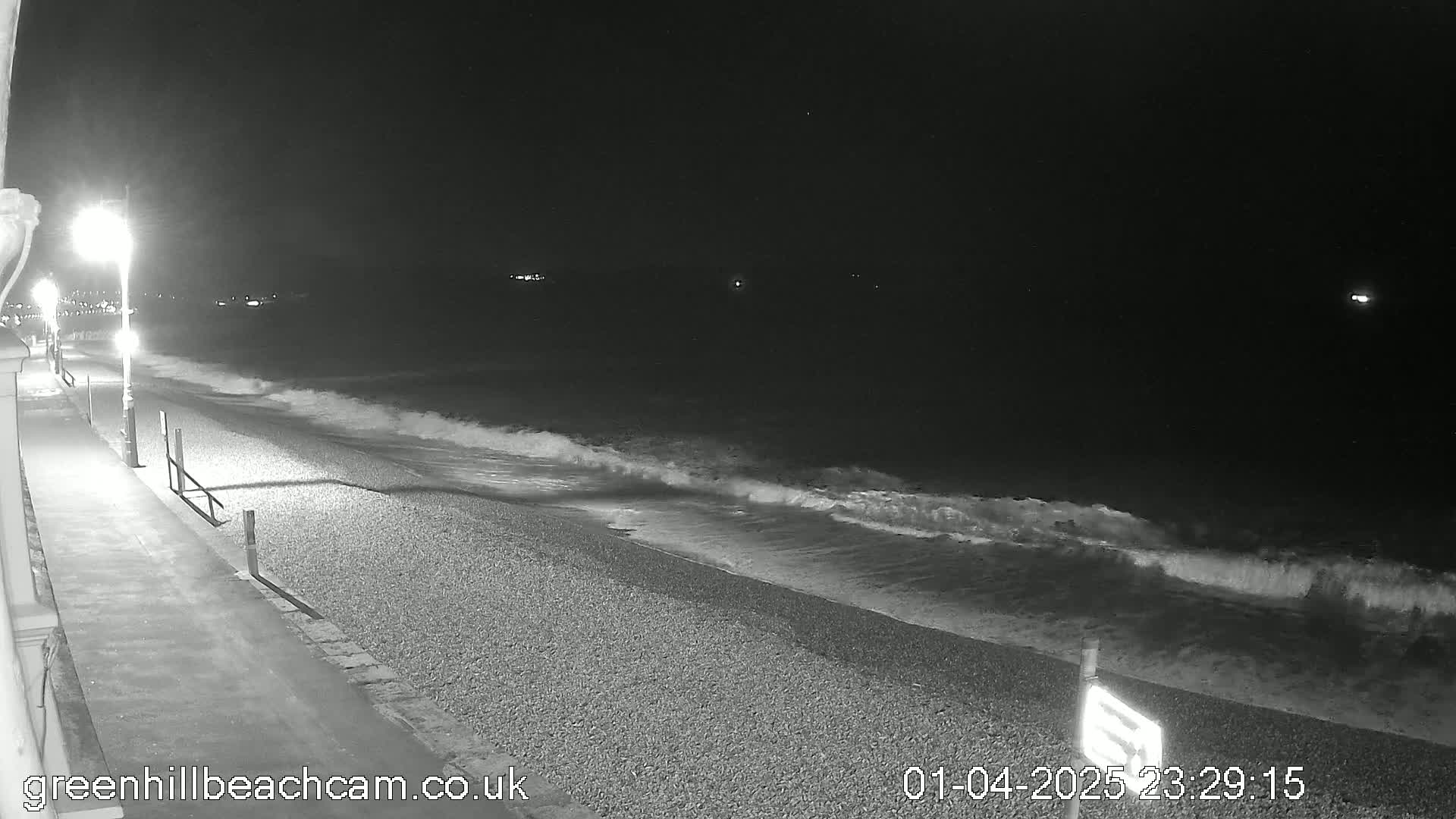 A nighttime view of a pebble beach with gentle waves lapping the shore, illuminated by streetlights along a paved walkway.