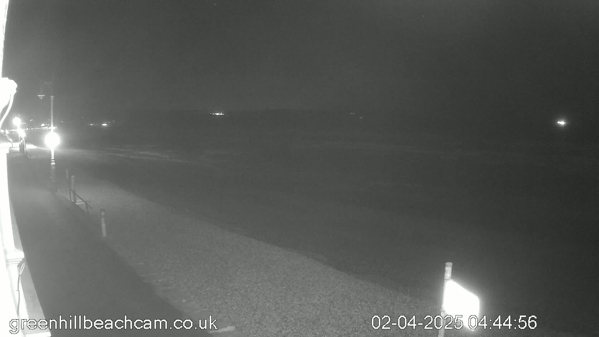 A nighttime view of a dark beach with a paved walkway, dimly lit by streetlights, and a calm sea in the background.