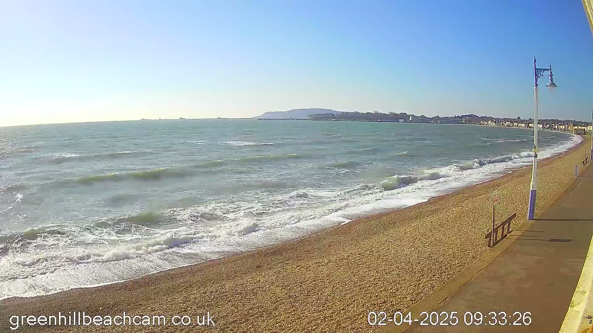 A beach with small waves lapping a pebble shore on a sunny day, with a town visible in the distance.