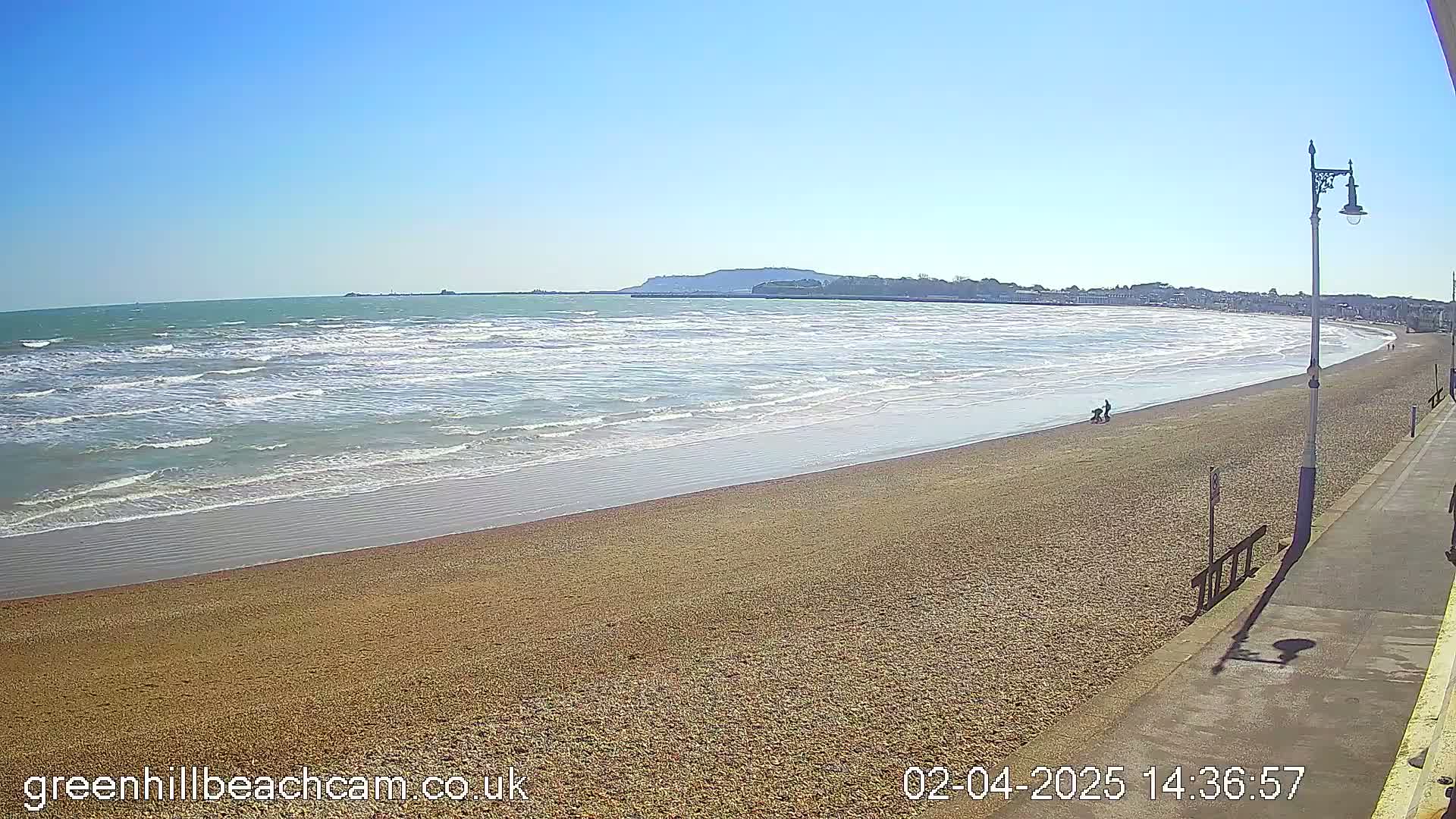 A sandy beach with small waves under a clear blue sky stretches to a distant coastline, where a few people are visible near the water's edge.