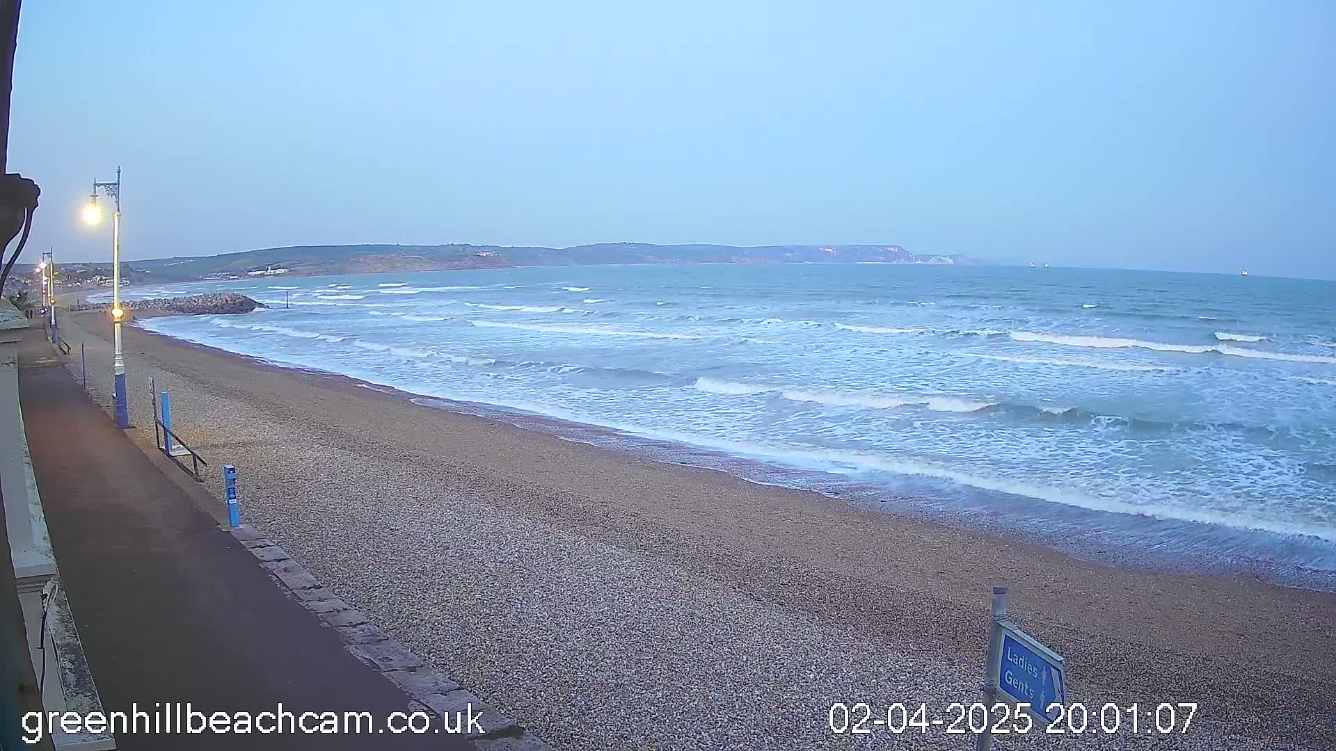 A pebble beach with moderate waves under a twilight sky stretches to a distant headland.