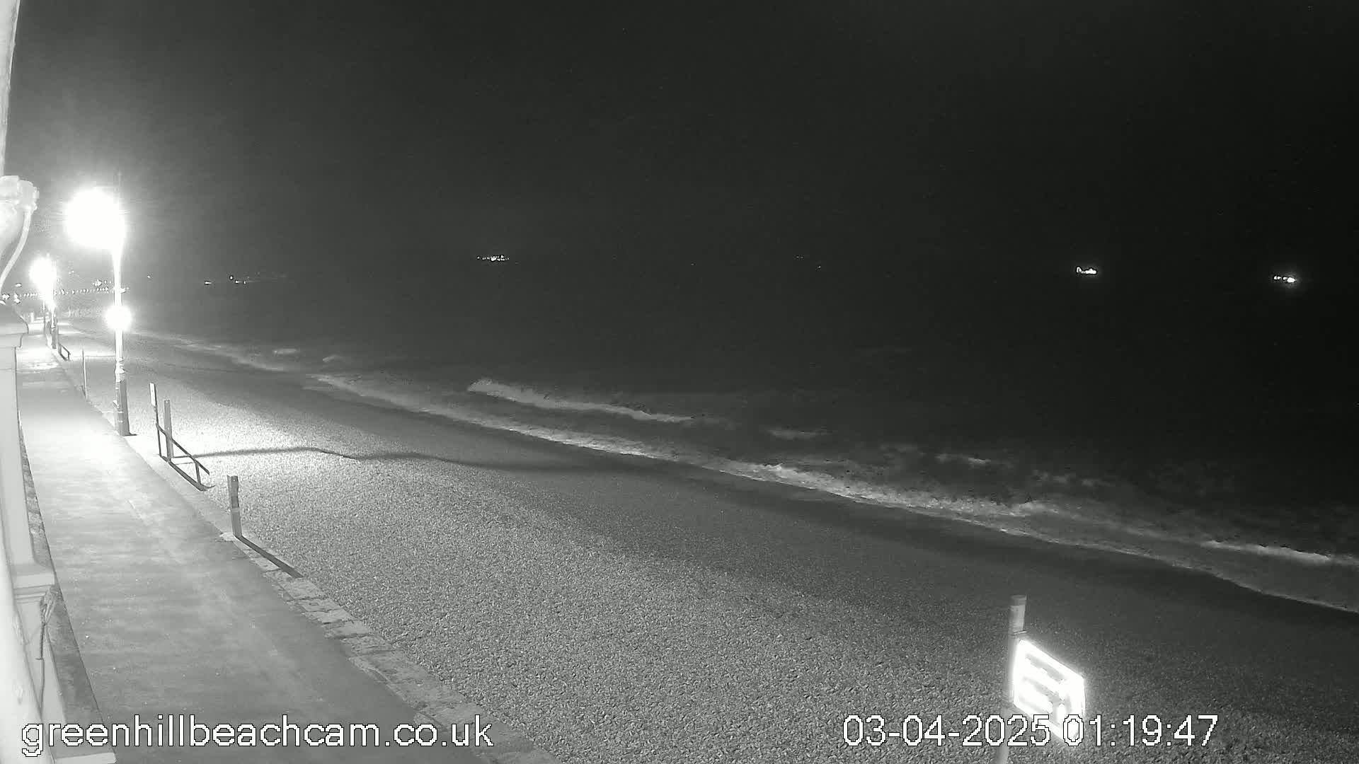 A pebble beach at night is gently lapped by small waves under a dark sky lit by several streetlights.
