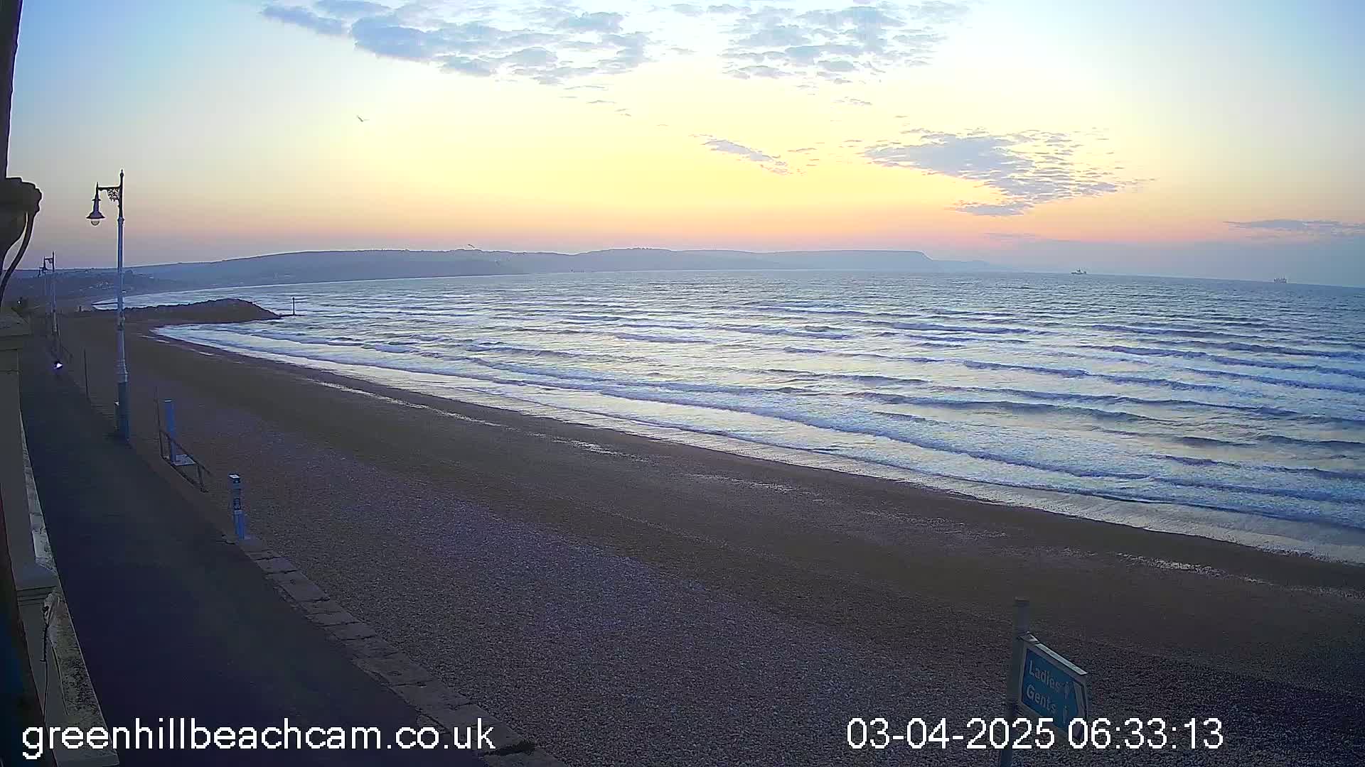 A pebble beach at sunrise, with gentle waves rolling onto the shore under a mostly clear sky.
