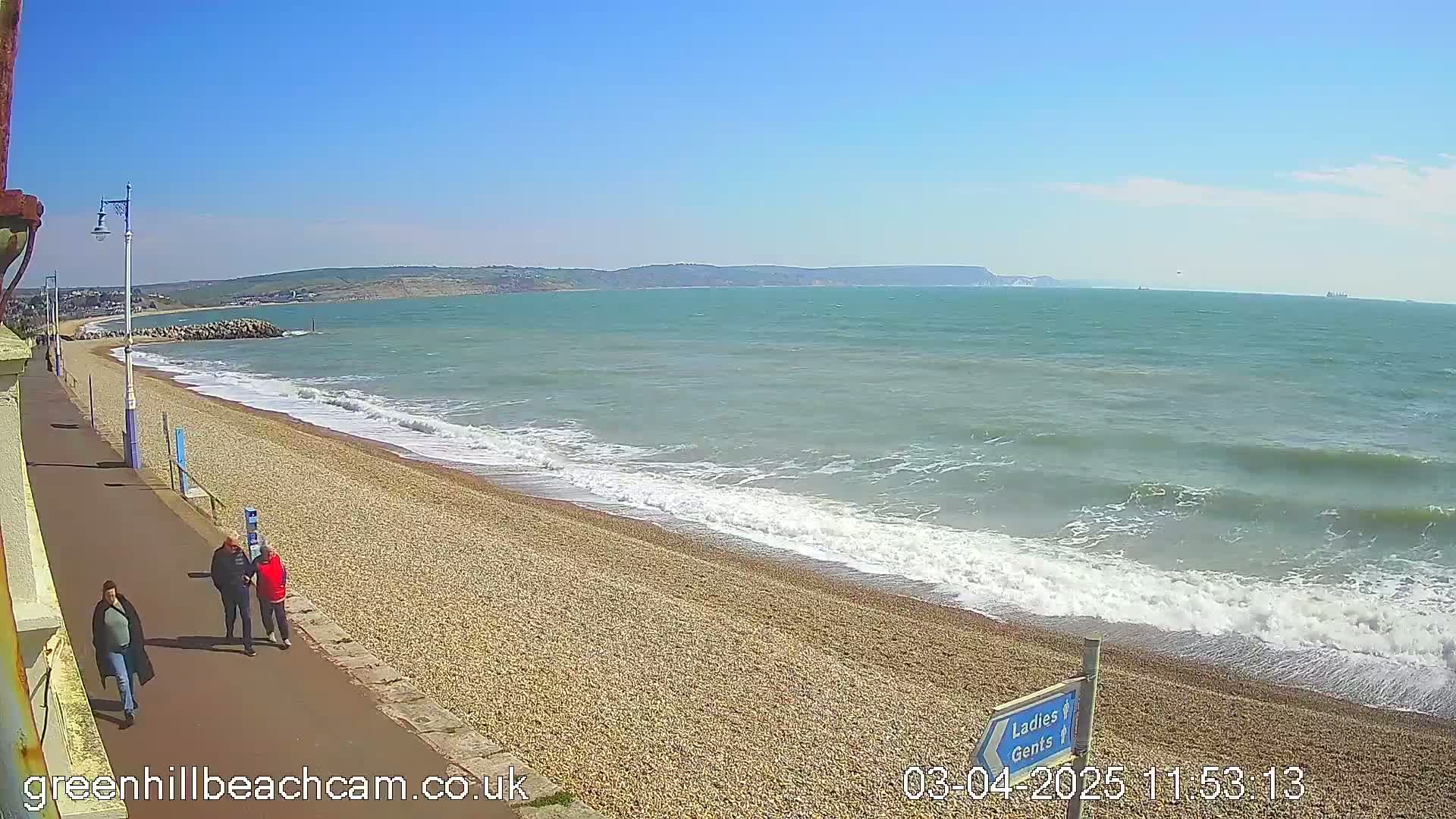 A pebble beach with moderate waves under a clear blue sky, and three people walking along a paved path beside it.