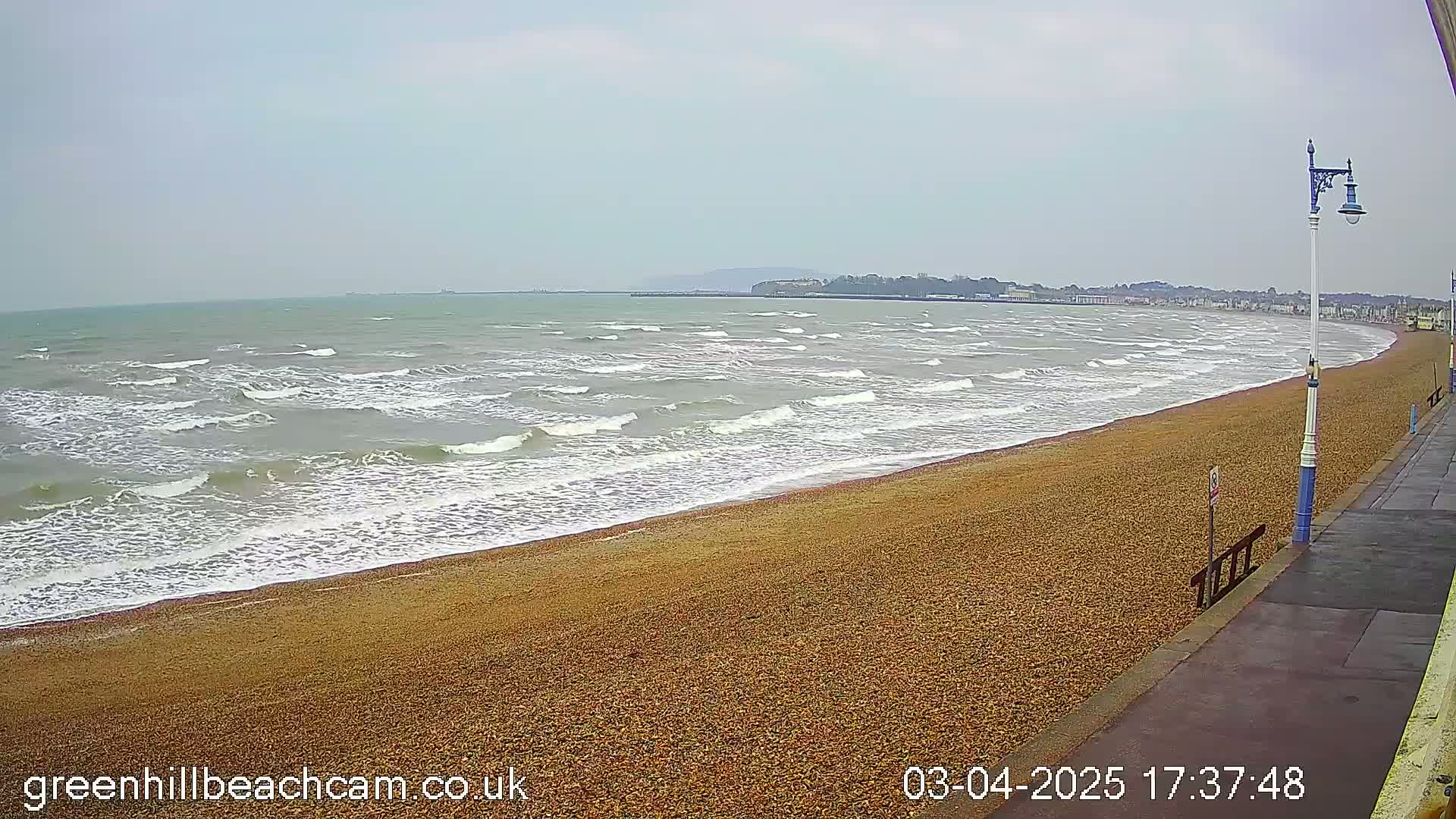A wide, windy beach with small waves lapping onto the shore, extending to a town visible in the distance under a cloudy sky.