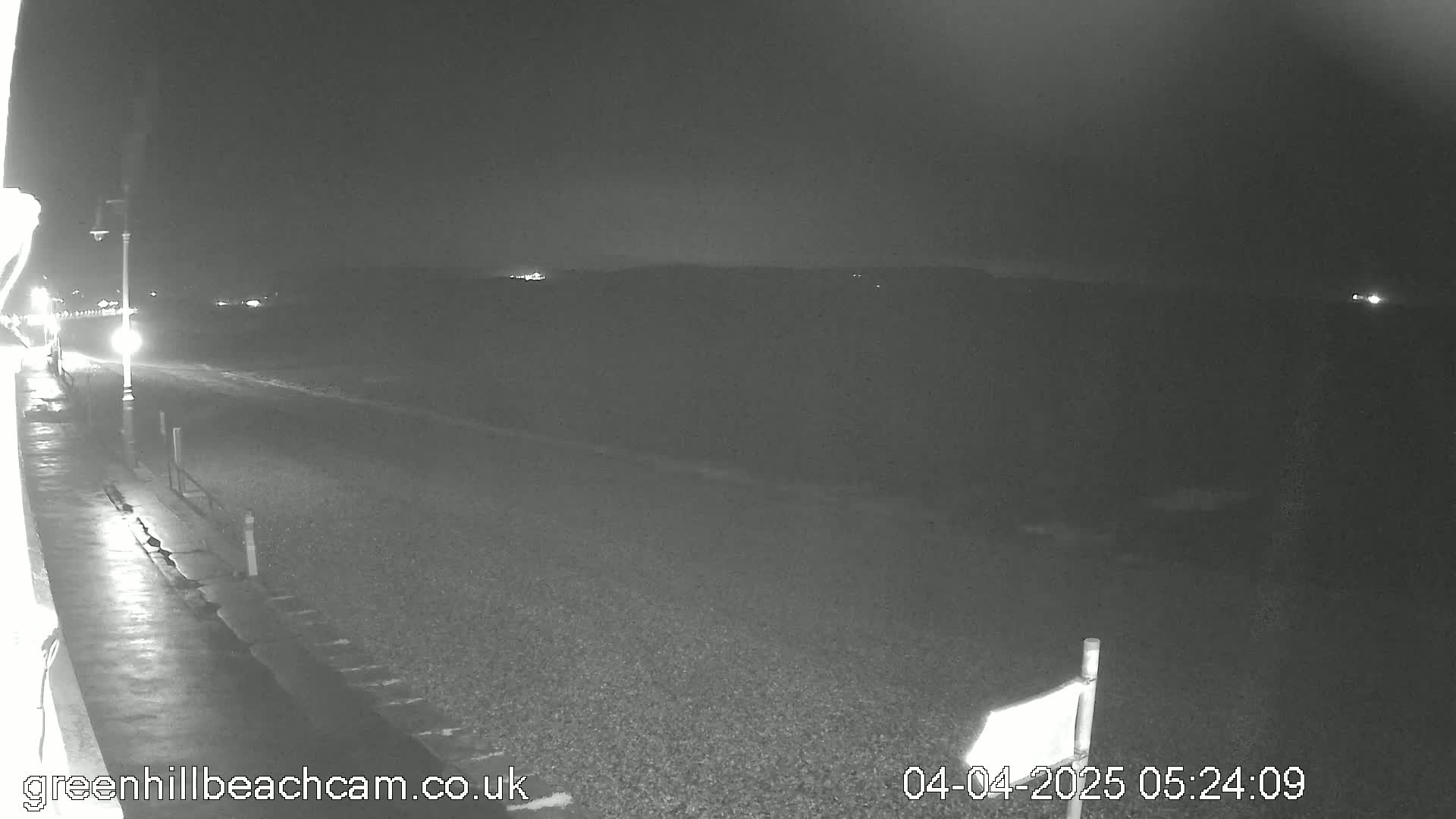 A nighttime, low-light view of a dark, pebbled beach and a paved walkway, with a few distant lights visible.