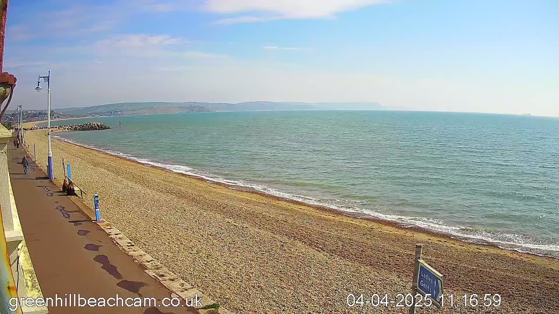 A pebble beach stretches along a calm, blue sea under a sunny sky, with a paved walkway and a few people visible in the distance.
