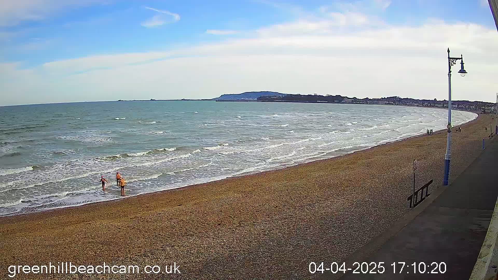 A sandy beach with small waves on a partly sunny day, a few people are visible on the beach and in the water near the shoreline.