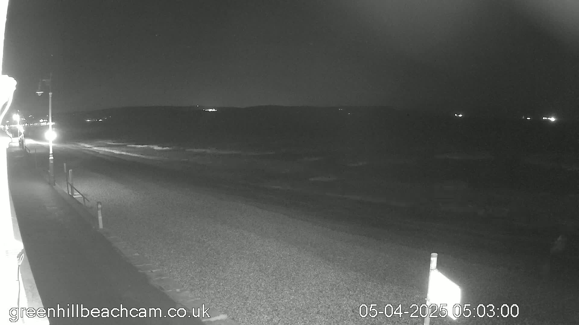 A nighttime, low-light view of a dark beach with a paved walkway and distant lights.