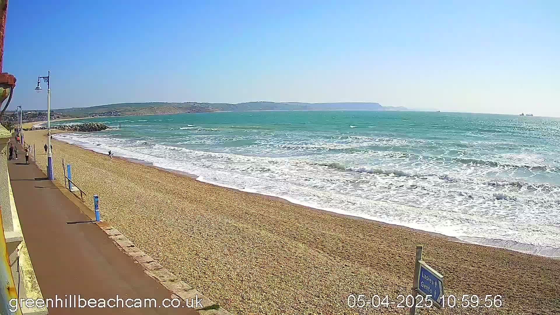 A pebbly beach with moderate waves under a clear blue sky, with a few people walking along the shore and promenade.