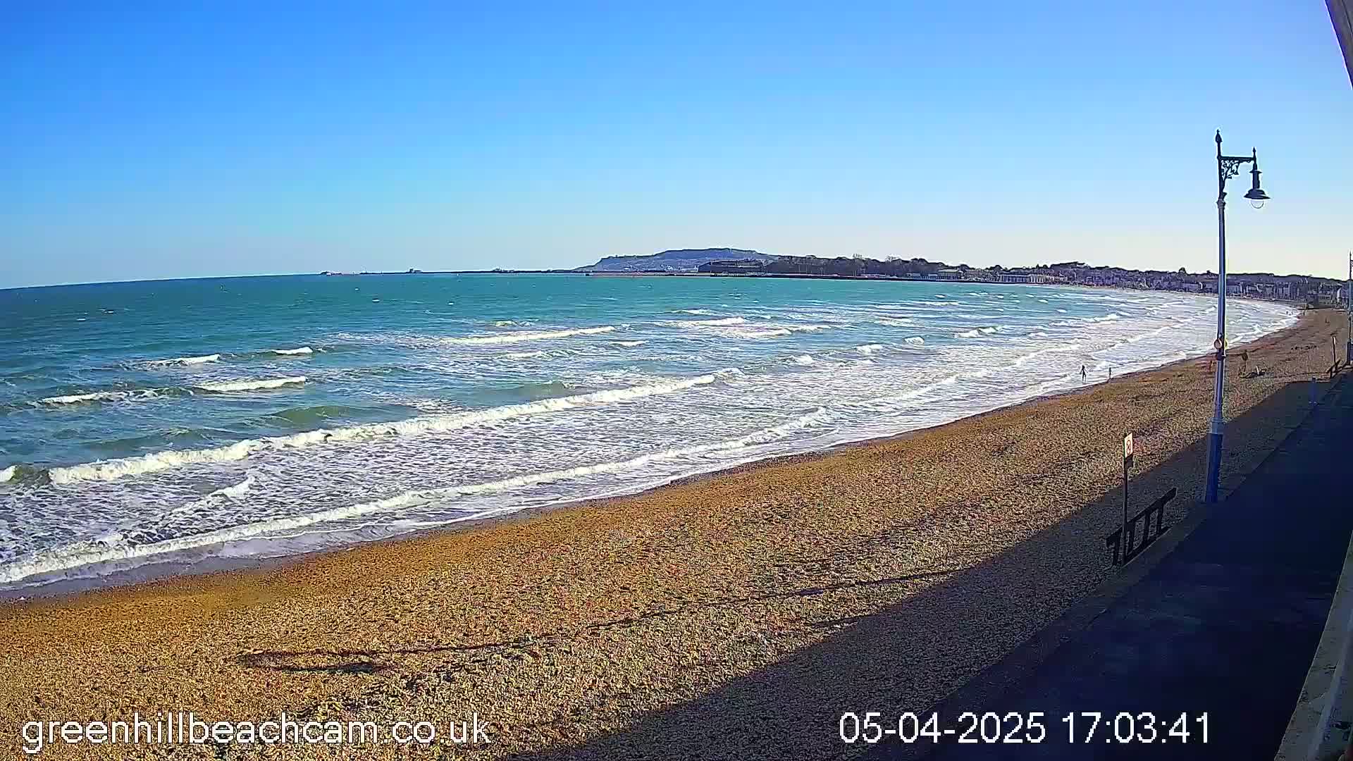 A sandy beach with gentle waves under a clear blue sky stretches toward a town and distant hills.