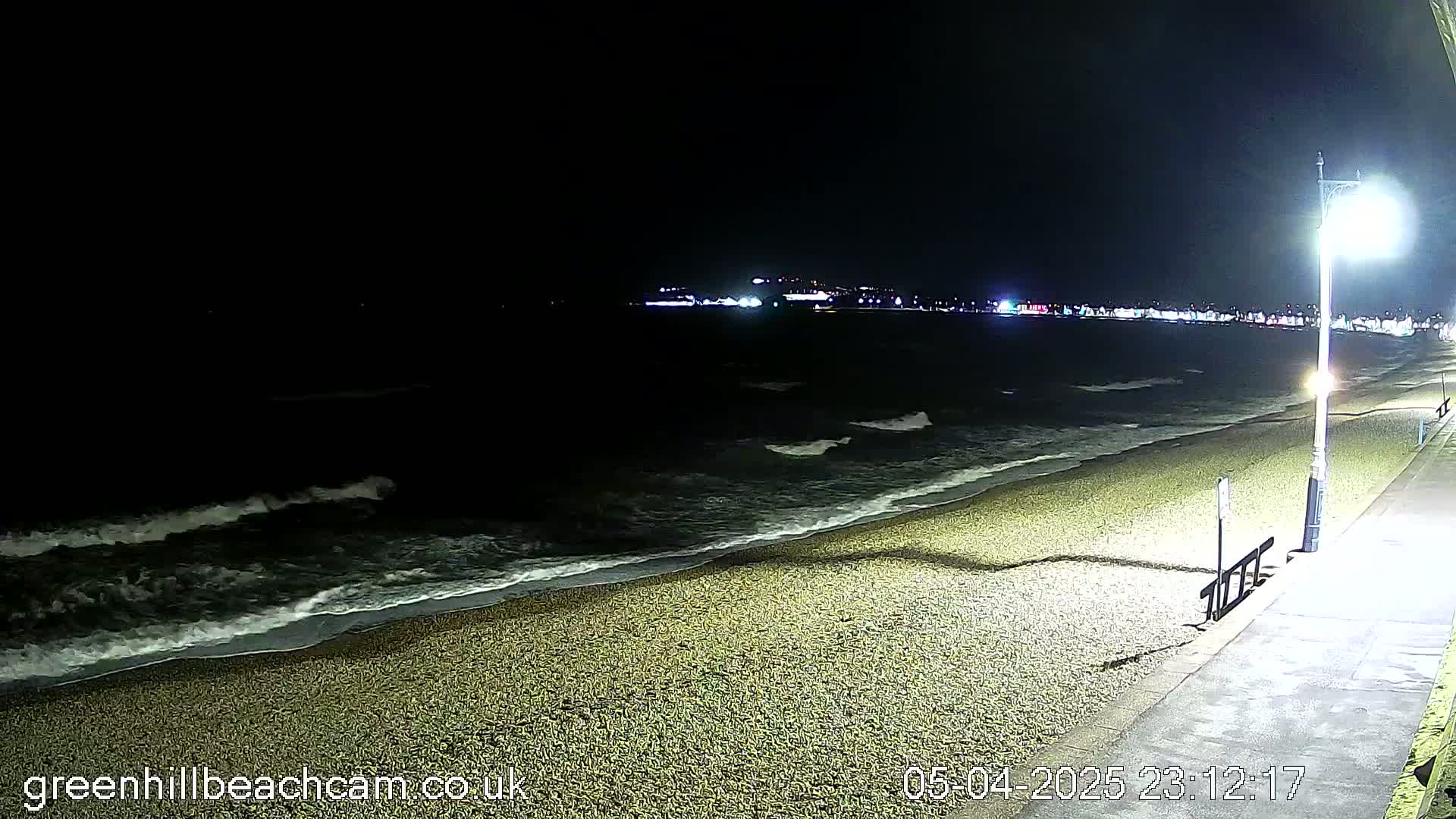 A dark, pebbled beach at night with gentle waves lapping the shore, illuminated by a streetlight and distant lights from a town.