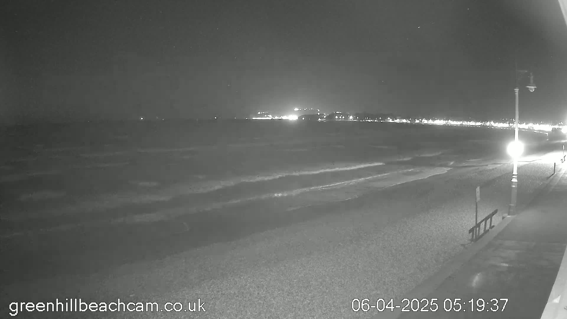 A dark, nighttime beach scene shows gently rolling waves lapping at a pebbled shore under a clear, starlit sky, with lights visible in the distance across the water.