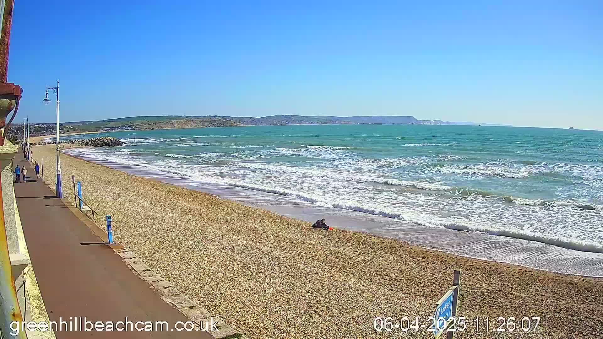 A pebble beach with moderate waves under a clear blue sky, and a few people walking along a paved walkway.
