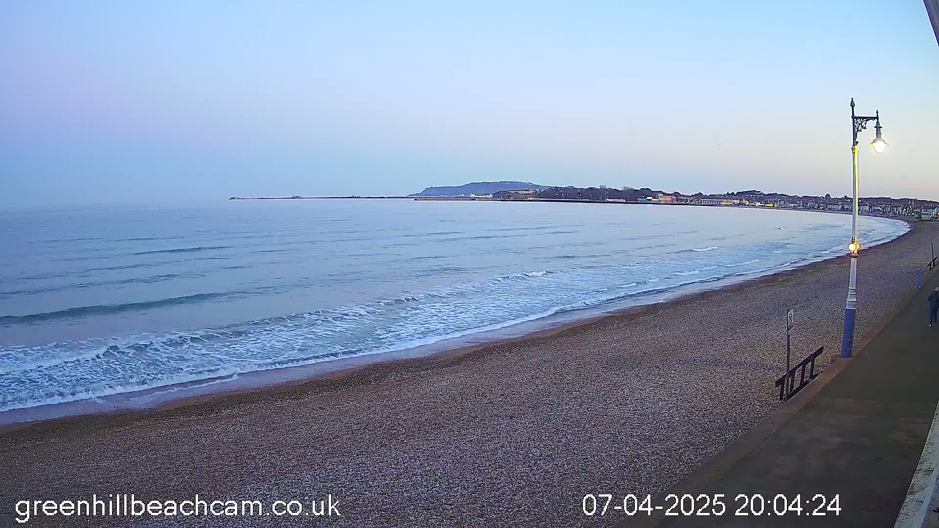 A long, pebbly beach slopes gently down to a calm, blue sea under a twilight sky.