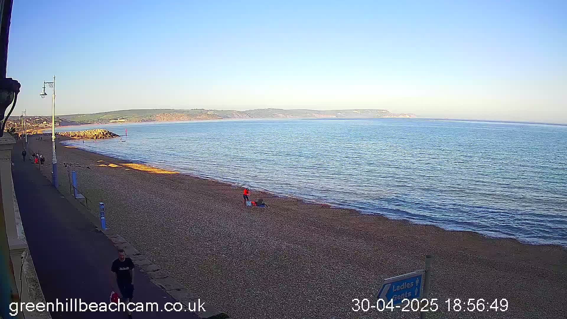 A pebble beach stretches along a calm, blue sea under a clear sky, with a few people walking on the beach and promenade.