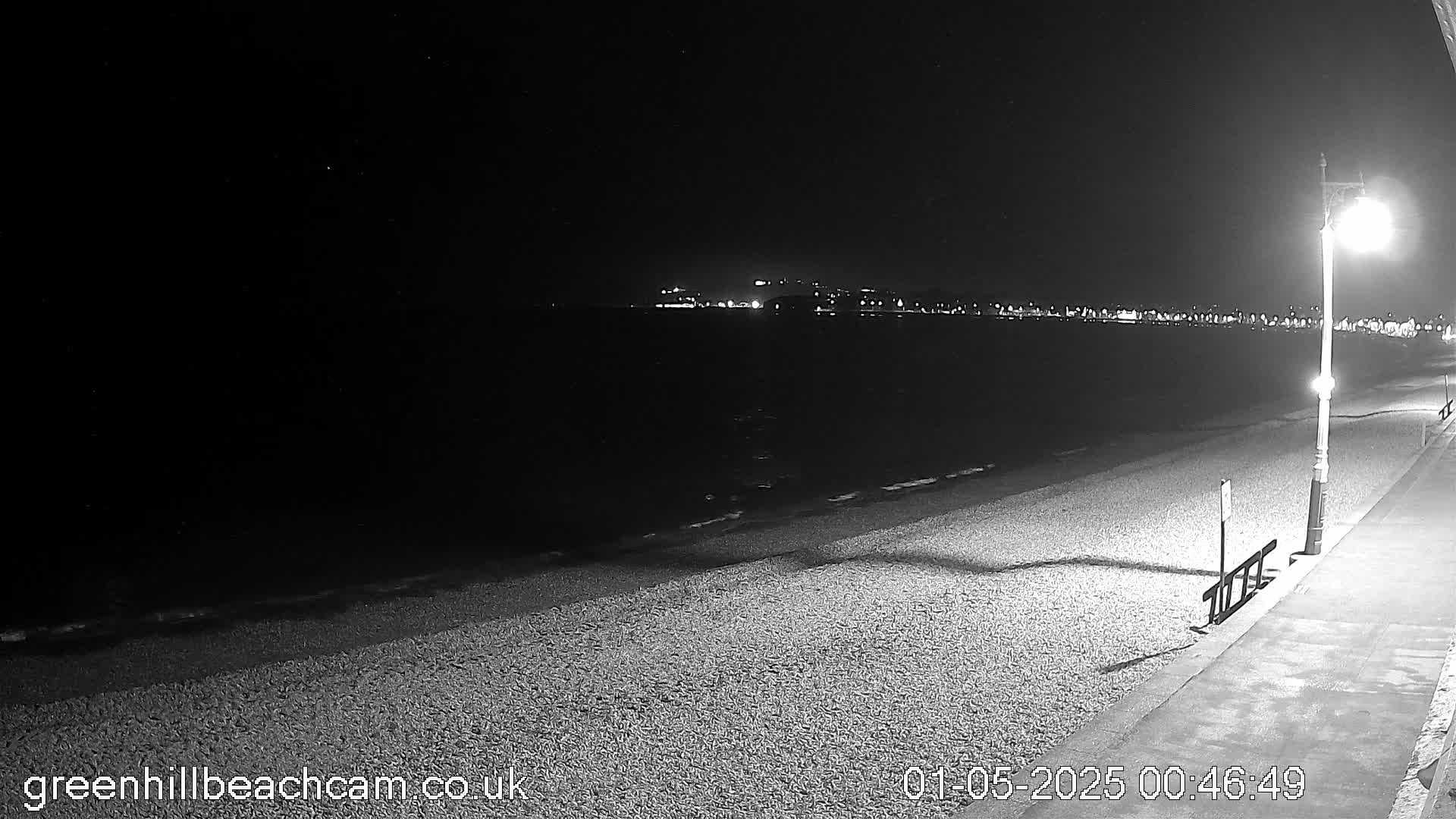 A dark, nighttime beach scene shows a pebble shore with a lamppost, a calm sea, and distant city lights.