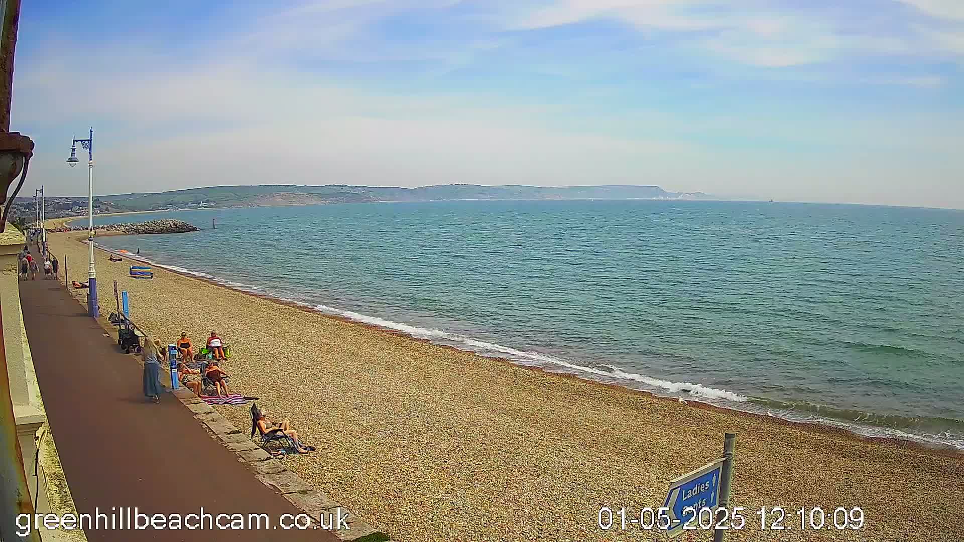 A pebble beach with a few people sunbathing stretches along a calm, blue sea under a mostly clear sky.