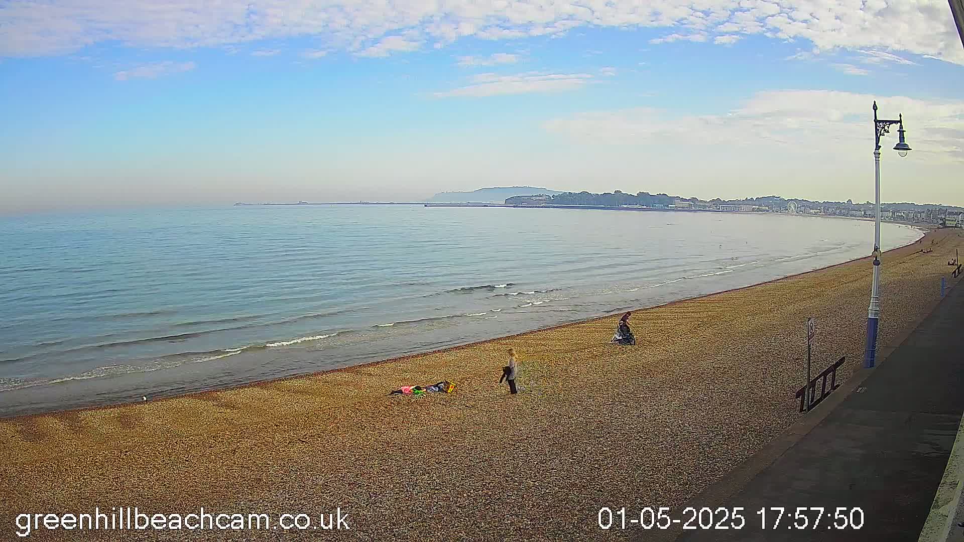 A mostly sunny day shows a long, gently sloping pebble beach with a few people, and calm waves lapping the shore, with a town visible in the distance.