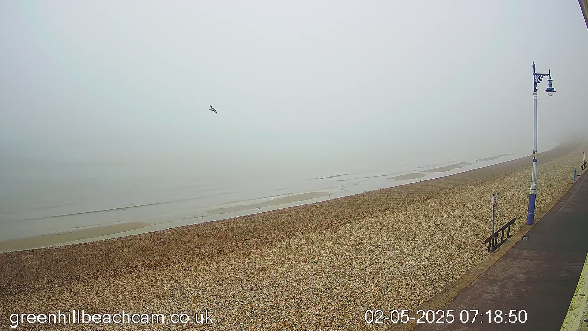 A foggy beach scene shows a wide, pebble-covered beach sloping down to a calm, gray sea under an overcast sky; a lamppost stands on a paved walkway overlooking the beach.