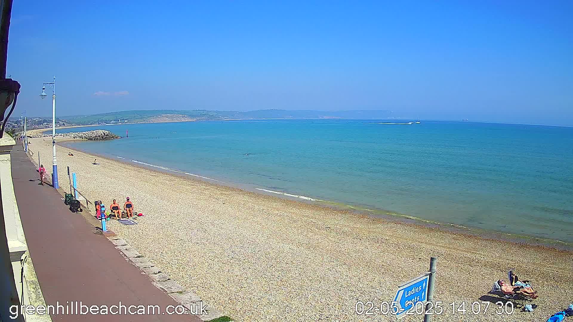 A pebble beach with a few people sunbathing stretches along a calm, clear blue sea under a bright, sunny sky.