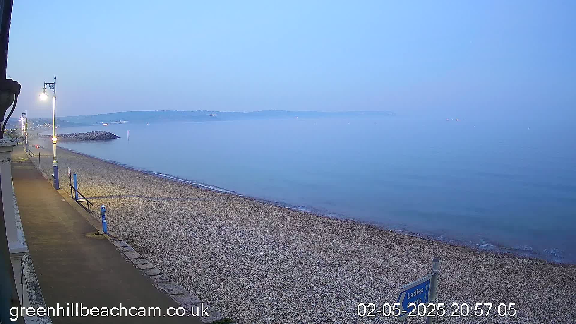 A hazy twilight scene shows a pebble beach and calm sea under a pale blue sky, with a walkway and lamps lining the shore.