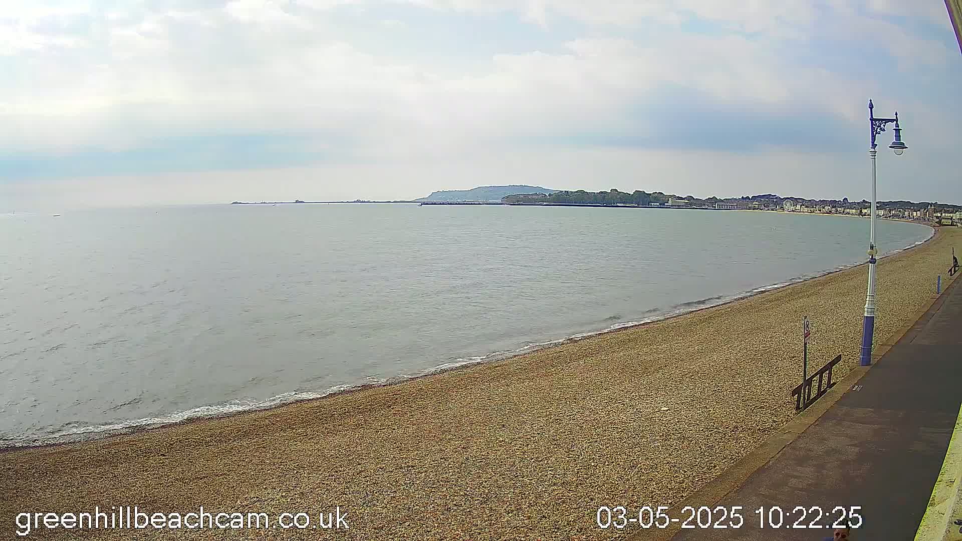 A pebble beach curves gently around a calm, grey sea under a mostly cloudy sky, with a distant town visible on the horizon.