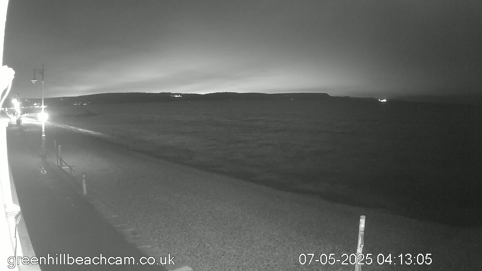 A nighttime, black and white view of a pebble beach and calm body of water with a distant shoreline under a dark, cloudy sky.