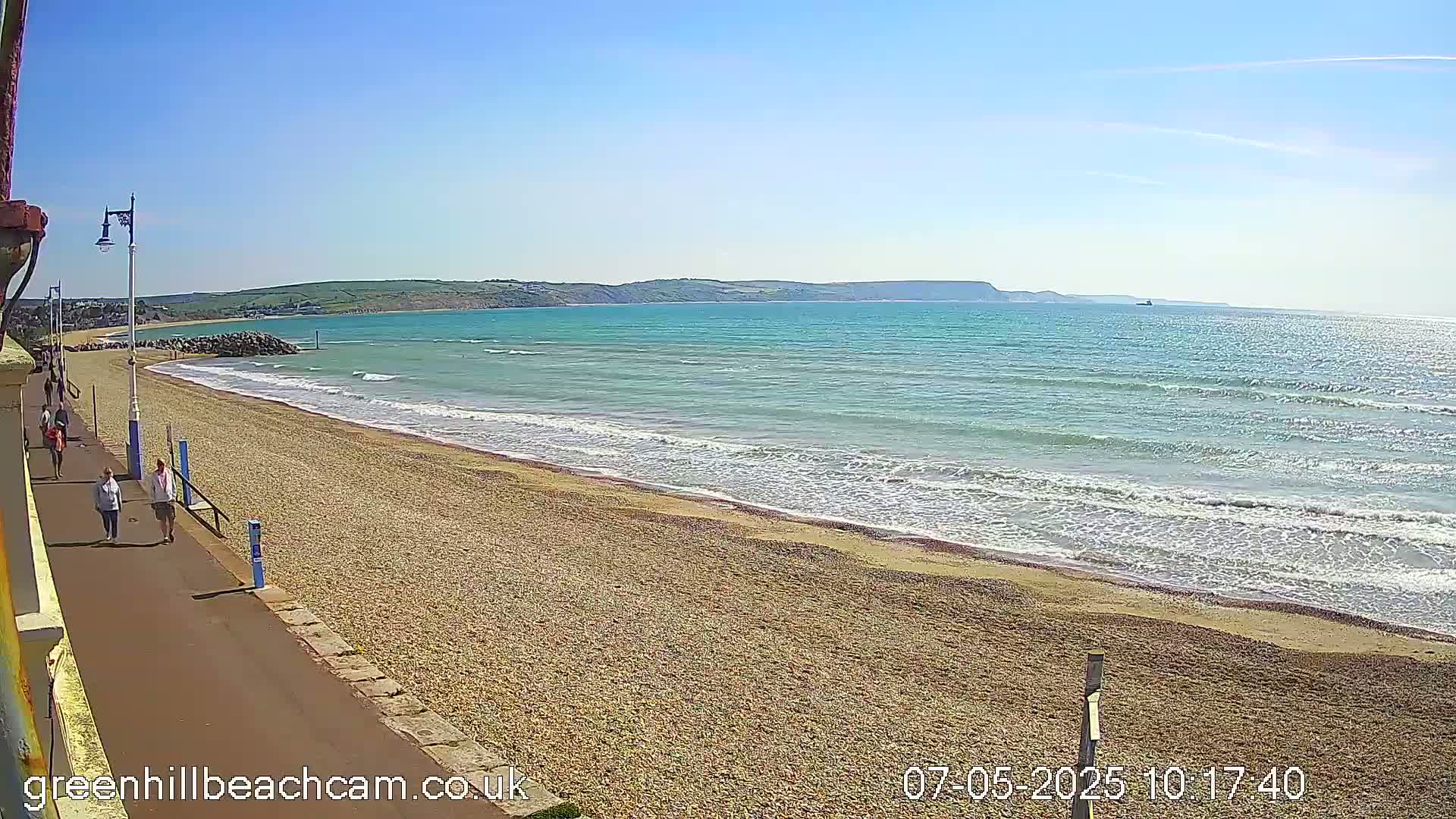 A pebble beach with a few people walking along a paved path beside it, gentle waves lapping the shore under a clear blue sky.