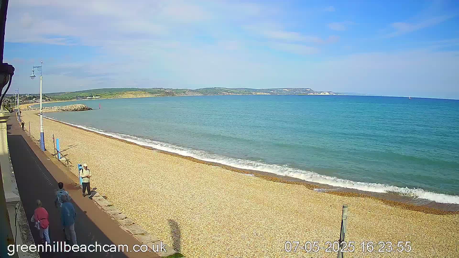 A pebble beach stretches along a calm, blue sea under a mostly sunny sky, with several people walking along the shore.