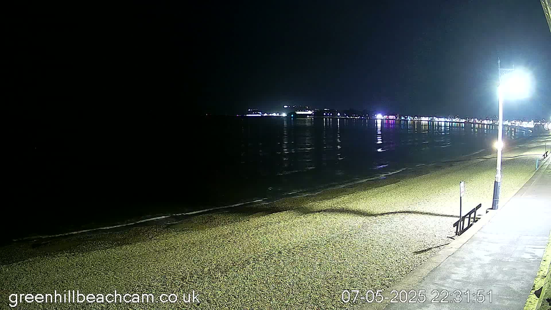 A dark, nighttime beach scene shows a calm, reflective body of water with lights across the horizon, a pebbled shoreline, and a paved walkway with a lamppost.