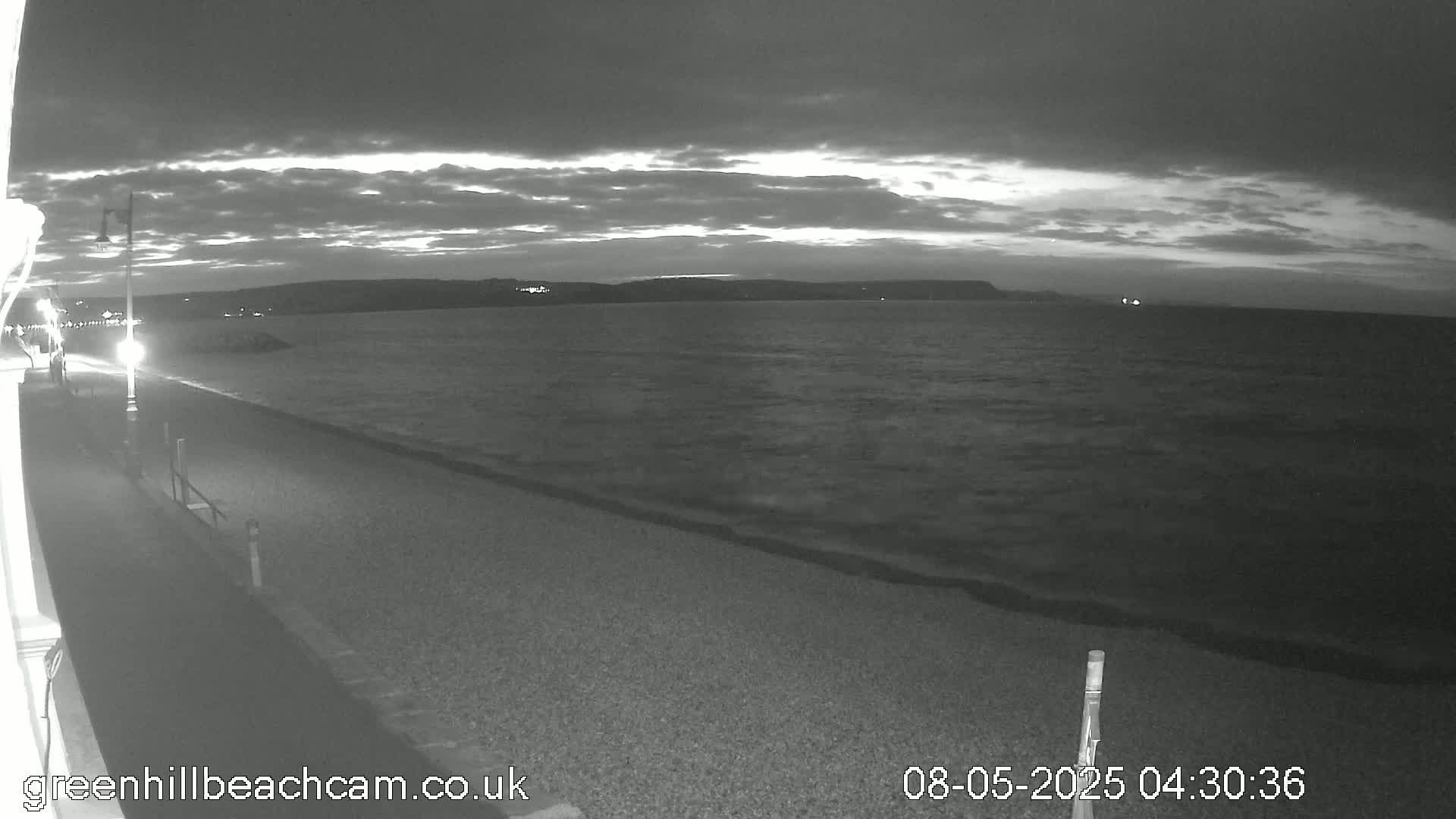 A dark, cloudy evening scene shows a calm sea lapping a pebble beach, with a paved walkway and a single lamppost visible in the foreground.